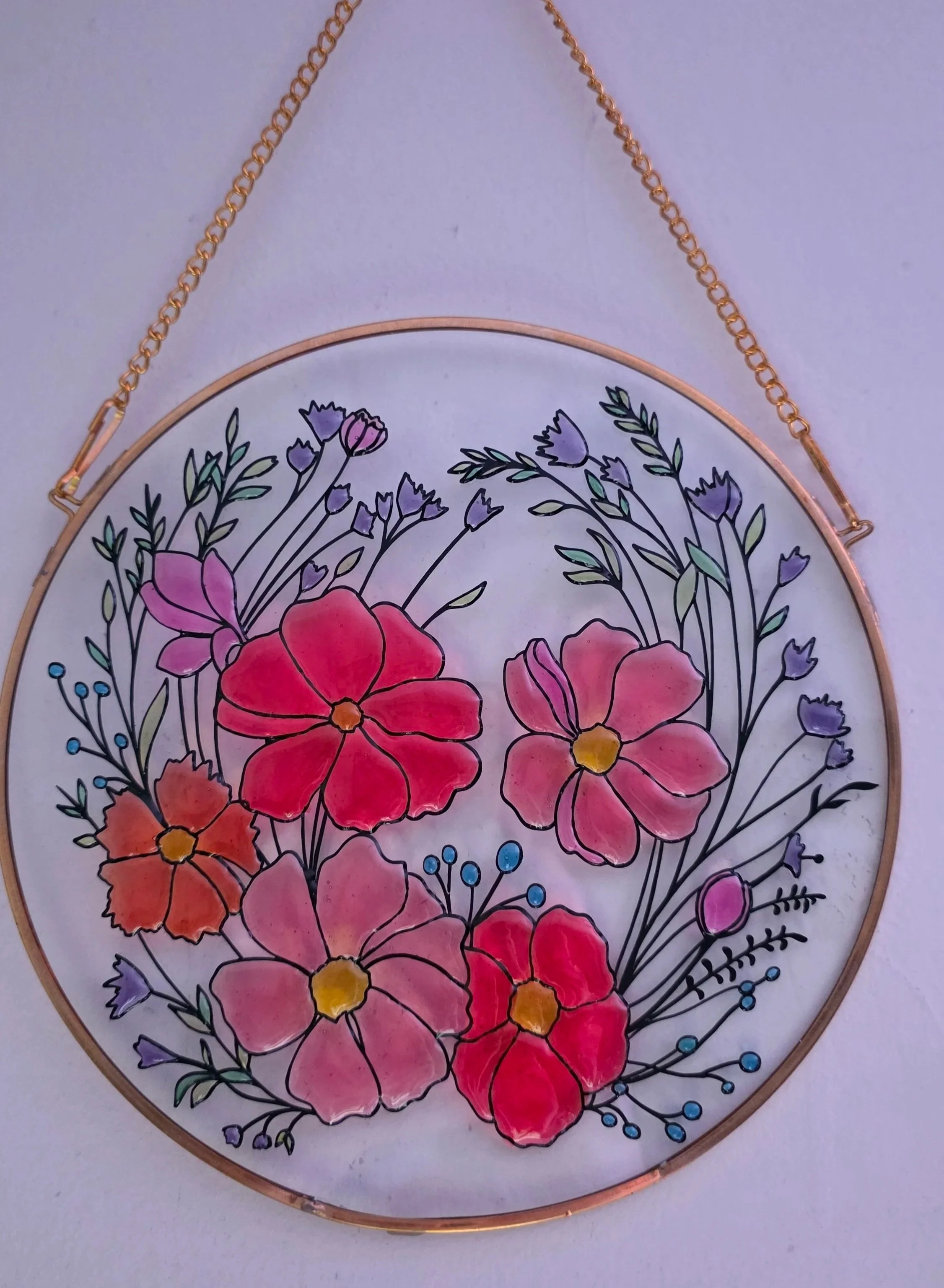 Colorful floral stained glass artwork framed in a round gold frame, hanging on a wall with a small blue hook and chain.