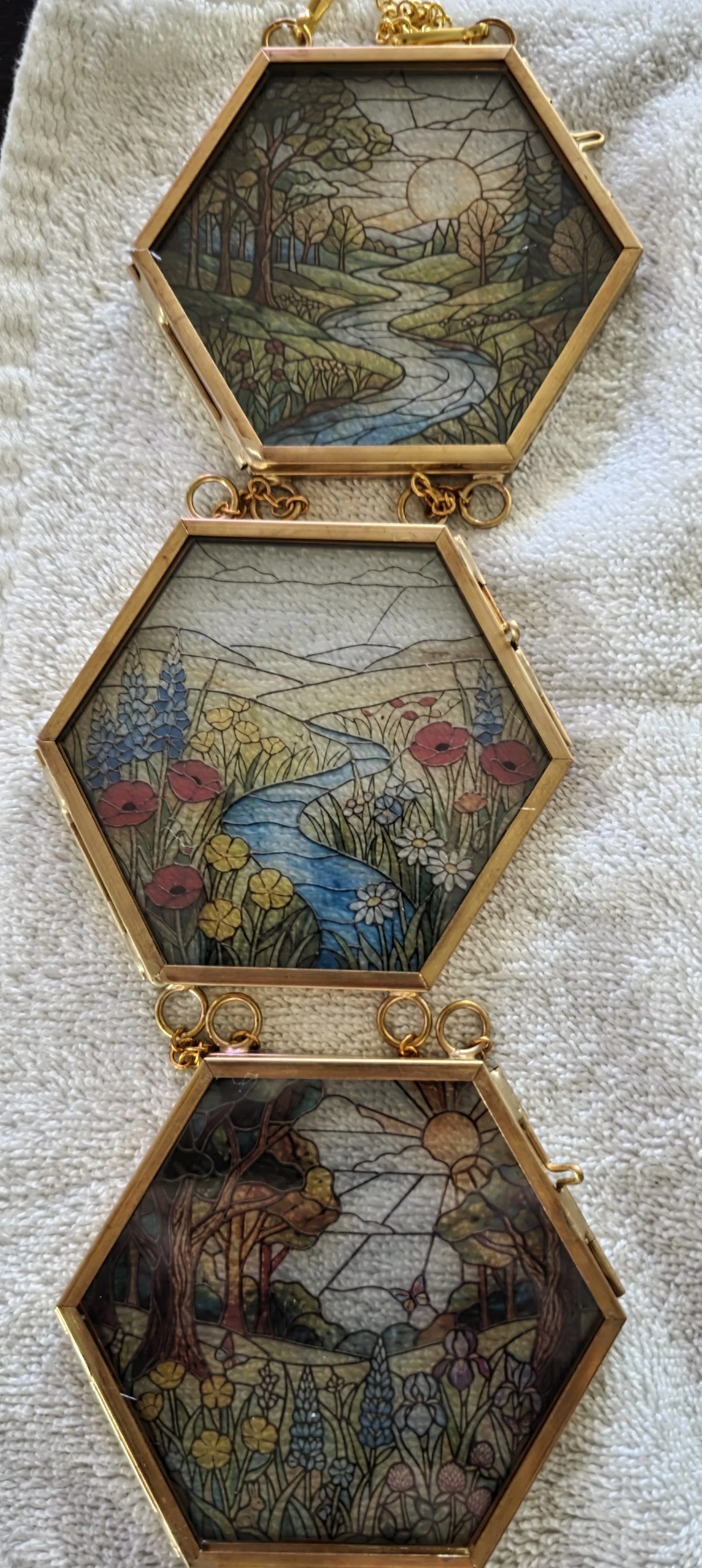 Three hexagonal glass frames with stained glass landscape scenes connected by small gold loops, resting on a textured fabric surface.