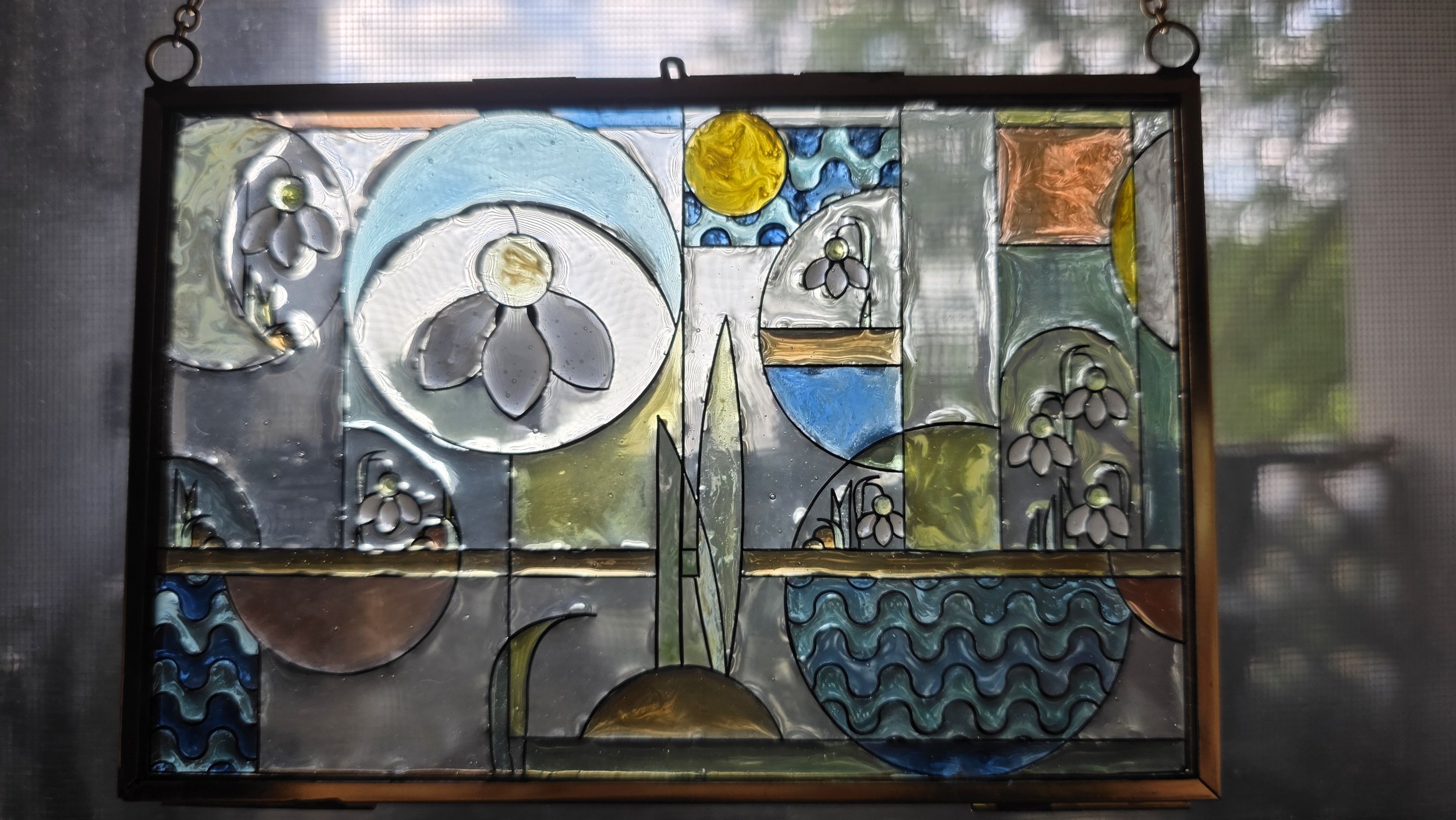 Colorful stained glass window featuring abstract floral designs, a yellow sun, and flowing wave patterns, framed in black.