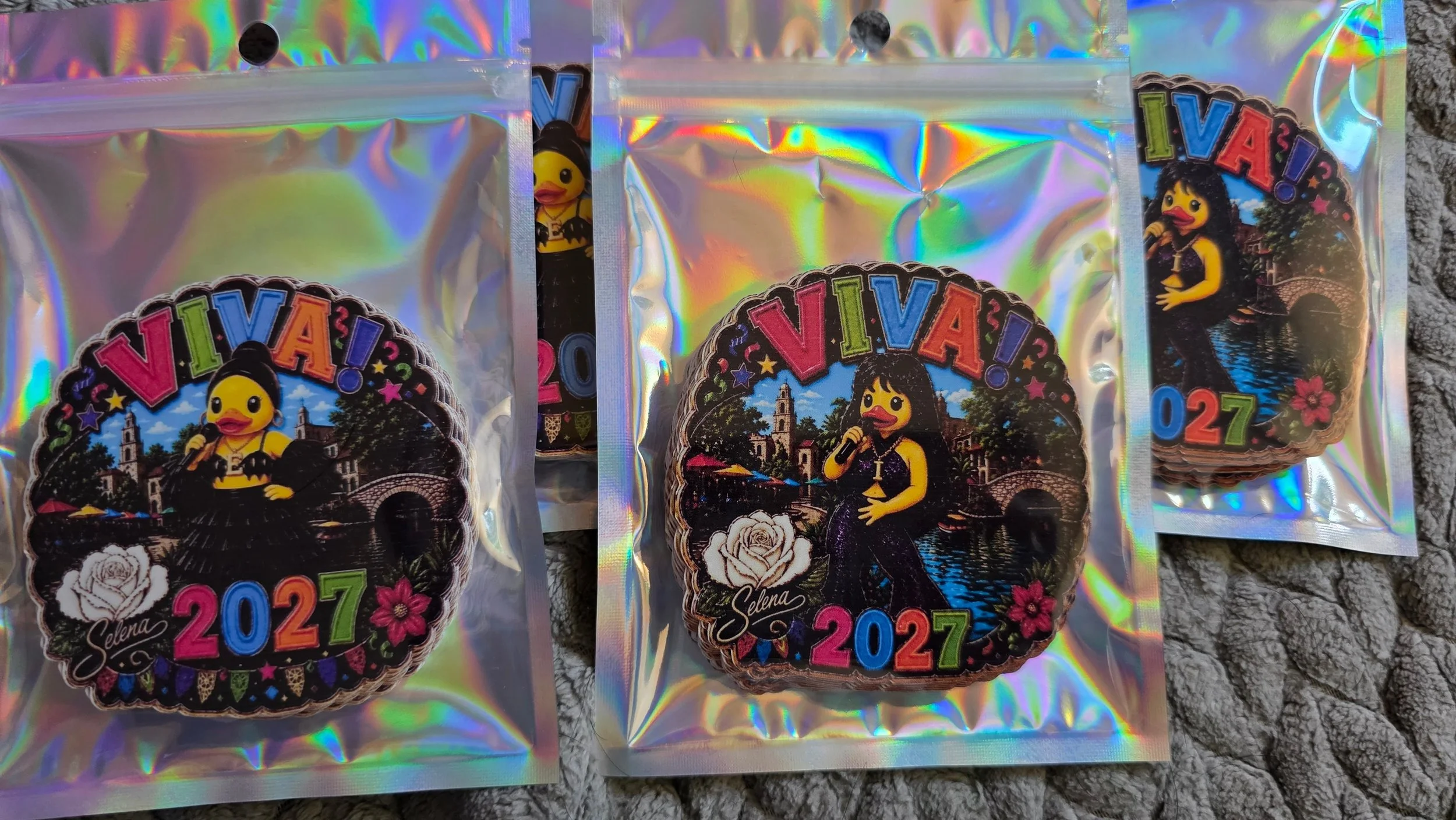 Three holographic party favor bags with colorful stickers displaying the year 2027, featuring a cartoon duck with a microphone, a woman named Selena holding a microphone, flowers, and a background of a cityscape with buildings and trees.