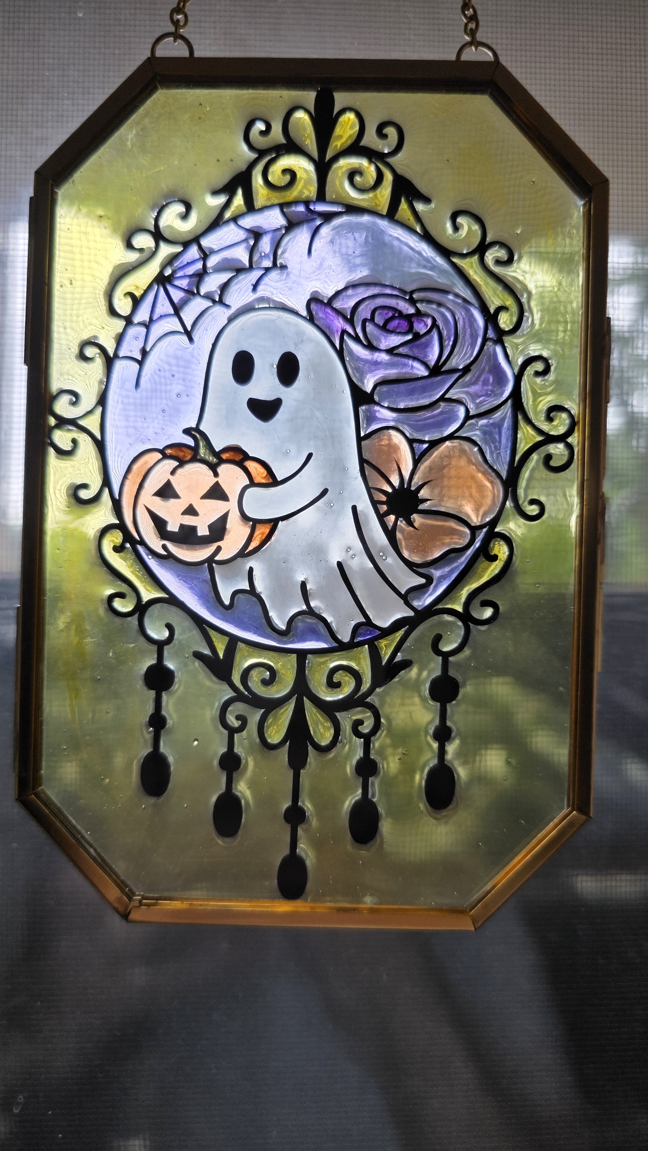 A decorative stained glass window featuring a cartoon ghost holding a carved pumpkin, with purple roses, a spider web, and hanging black chains with spider accents.