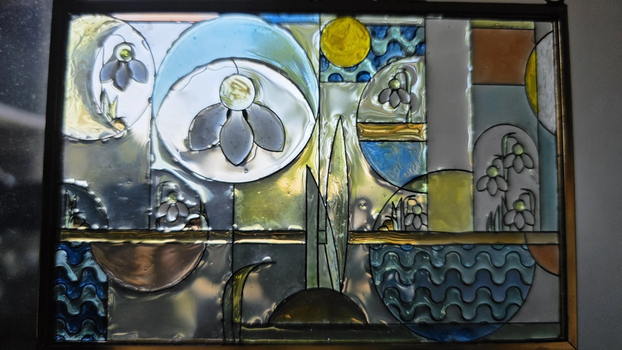 A colorful stained glass window with abstract floral and geometric patterns, featuring flowers, circles, and wavy lines in blue, yellow, green, gray, and brown.