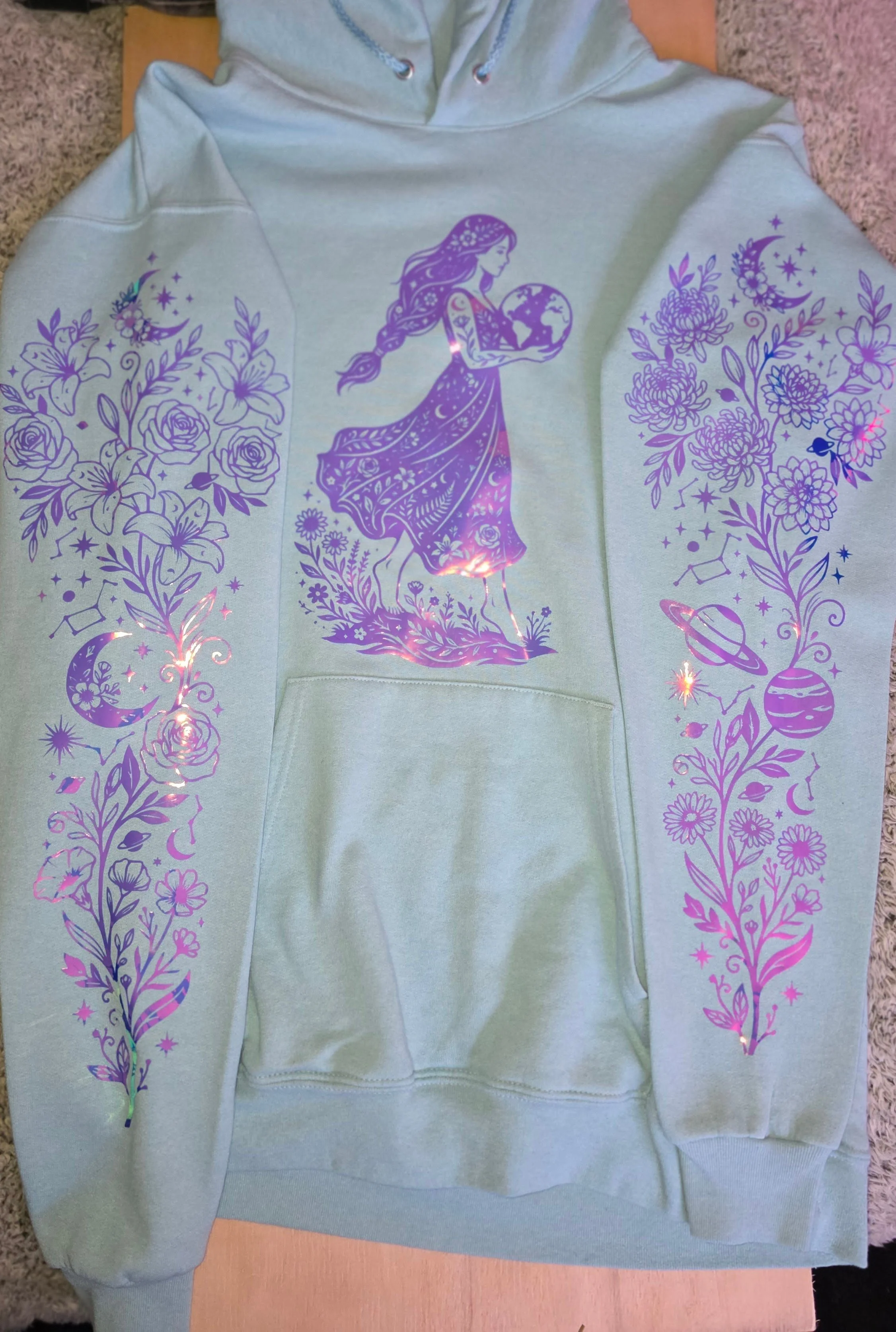 Light-colored hoodie with a purple illustration of a woman holding the Earth, surrounded by floral and celestial designs on the sleeves.