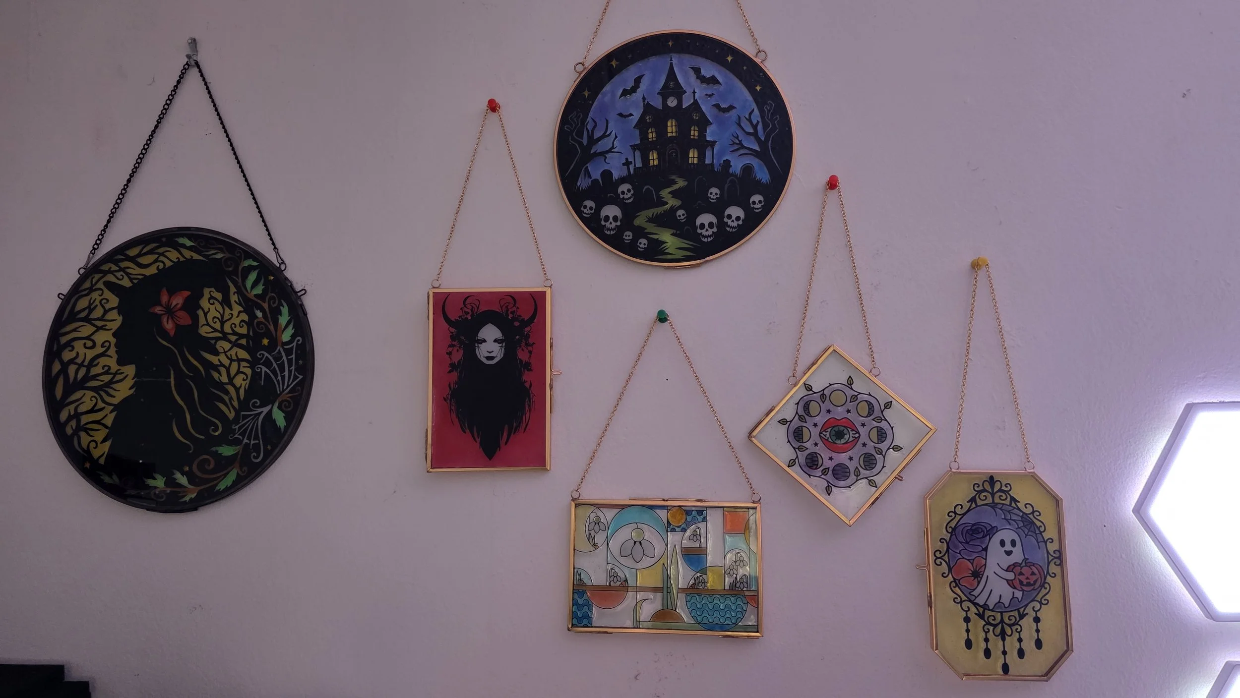 Wall decorated with various hanging art pieces, including a haunted house scene, a woman with long hair and horns, a ghost with a pumpkin, and abstract stained glass designs.