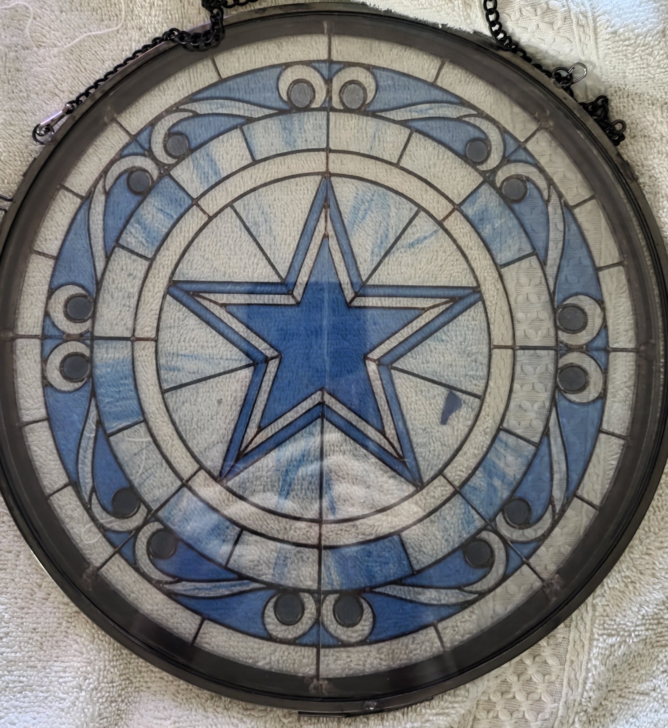 A round stained glass window featuring a blue and white star design with intricate black metal framing, placed on a textured white fabric surface.