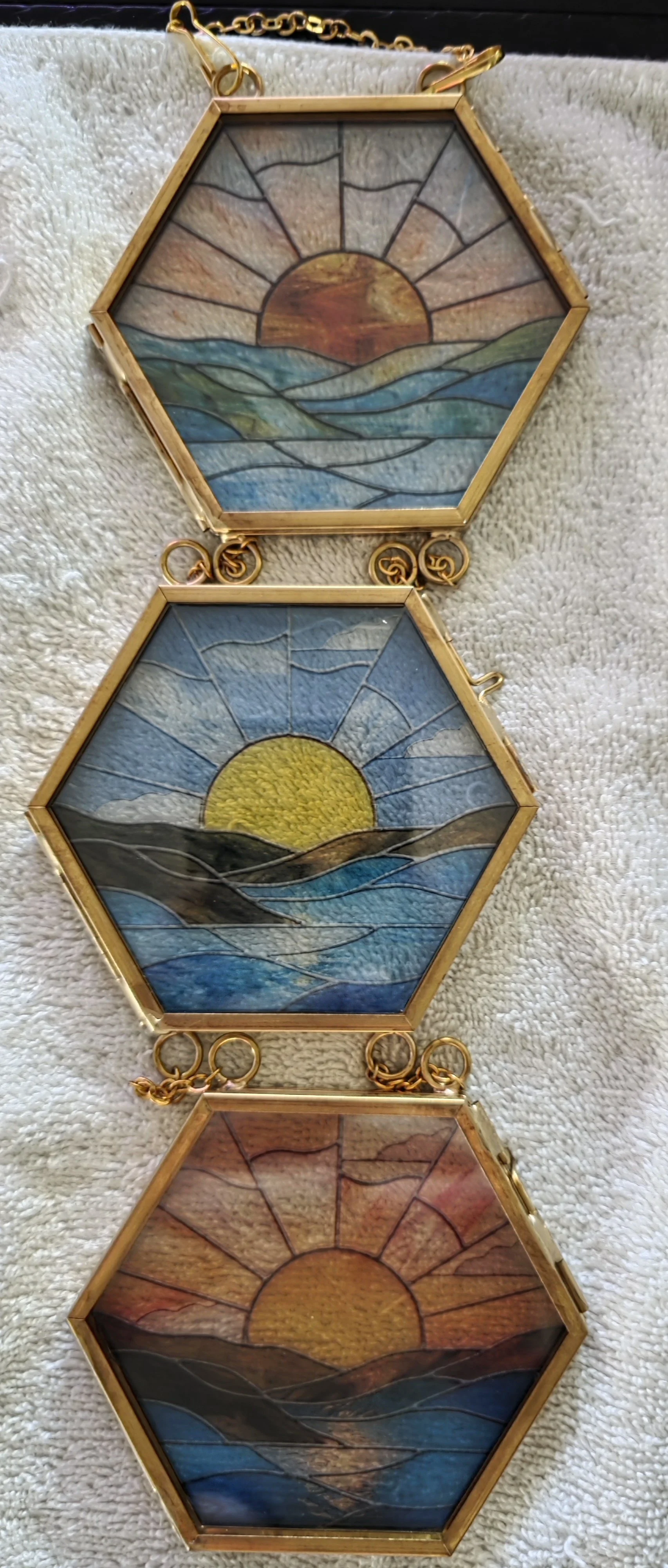 Three hexagonal stained glass pendant charms featuring sunrise and sunset scenes over mountains and water, with gold-colored metal frames and lobster clasp chains.