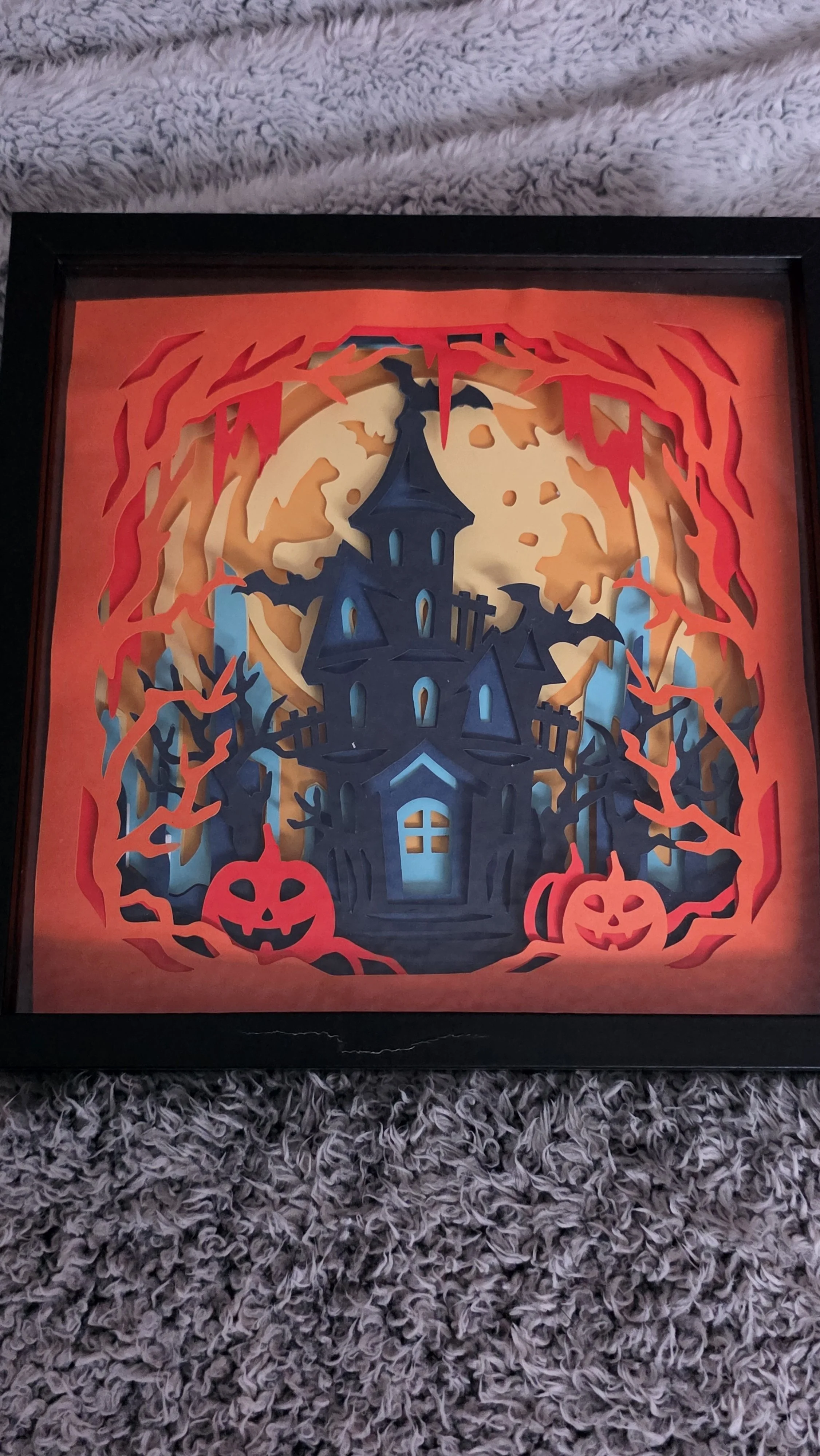 A Halloween-themed paper cutout artwork of a haunted house with a full moon, surrounded by red flames, purple trees, and two smiling pumpkin jack-o'-lanterns at the bottom.