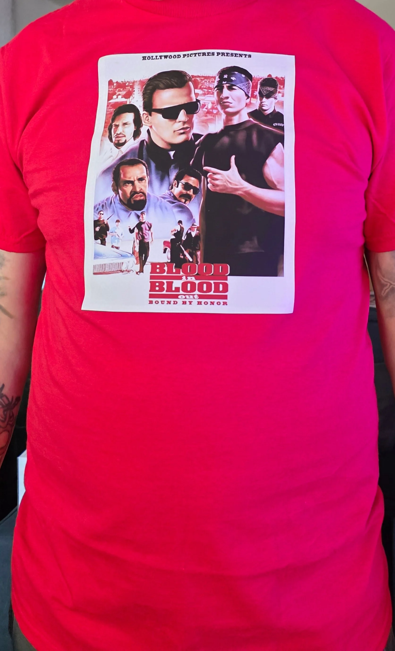A person wearing a red T-shirt with a graphic print of characters from the film "Blood in Blood Out".