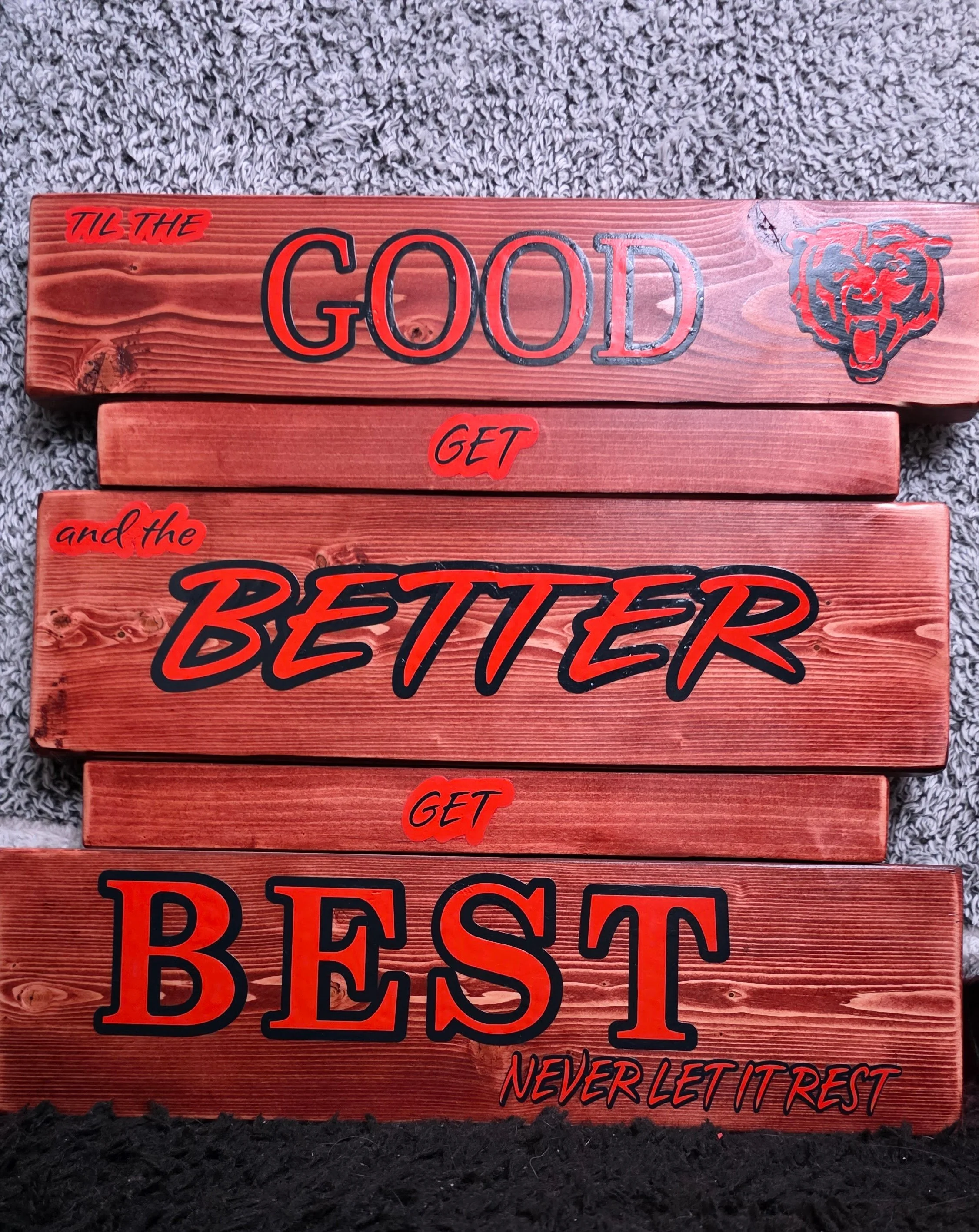 A wooden sign with text that says "Good Better Best, Never let it rest, till the good gets better and the better gets best" with the Chicago Bears logo. The sign is placed on dark soil or gravel.