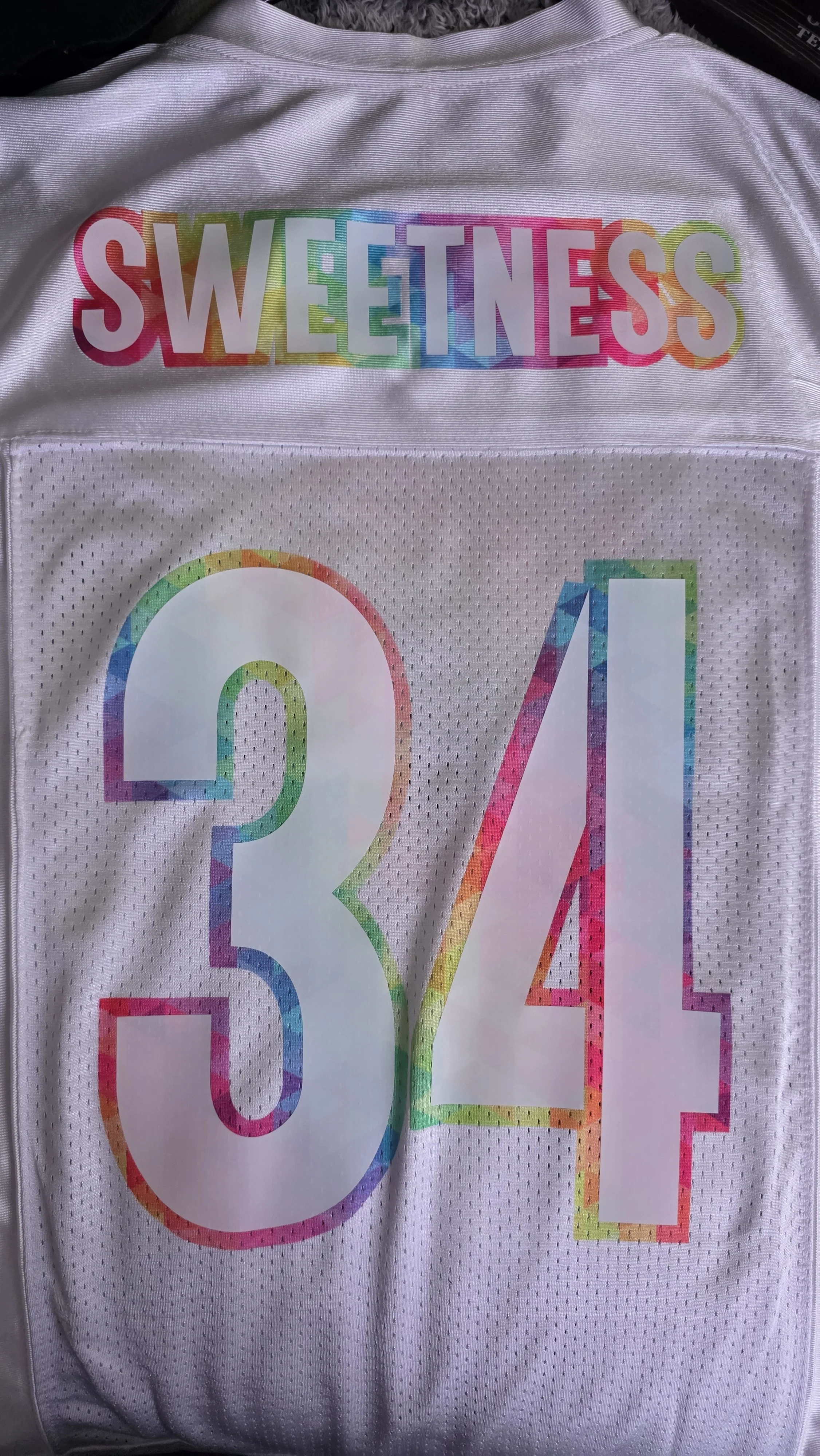 A white sports jersey with rainbow-colored large numbers 54, and a rainbow word 'SWEETNESS' at the top.