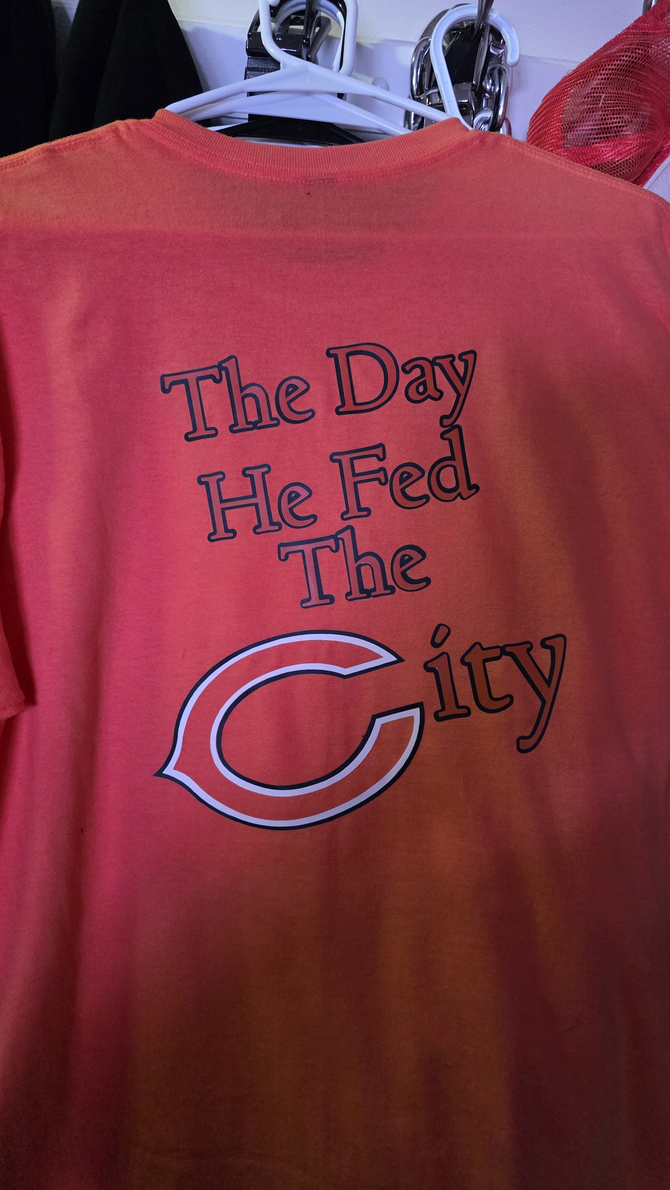 A red T-shirt hanging on a hanger with text that reads, 'The Day He Fed The City,' and a large letter 'C' logo.