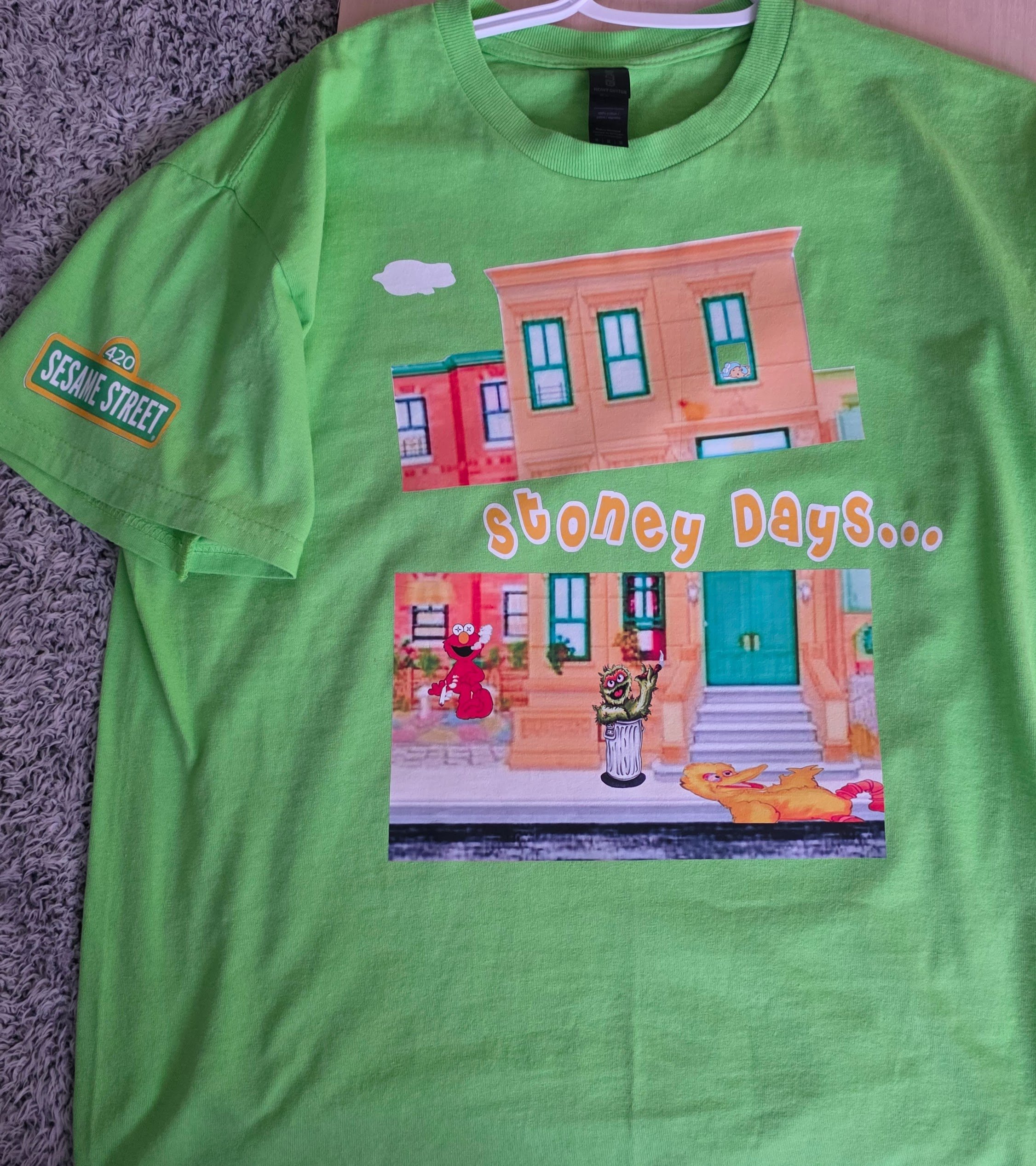 Bright green T-shirt lying on a textured gray surface. It features a cartoon illustration of Sesame Street characters on a brown building facade with green window frames, a Sesame Street sign, and the words "Stony Days" in orange and white. The shirt