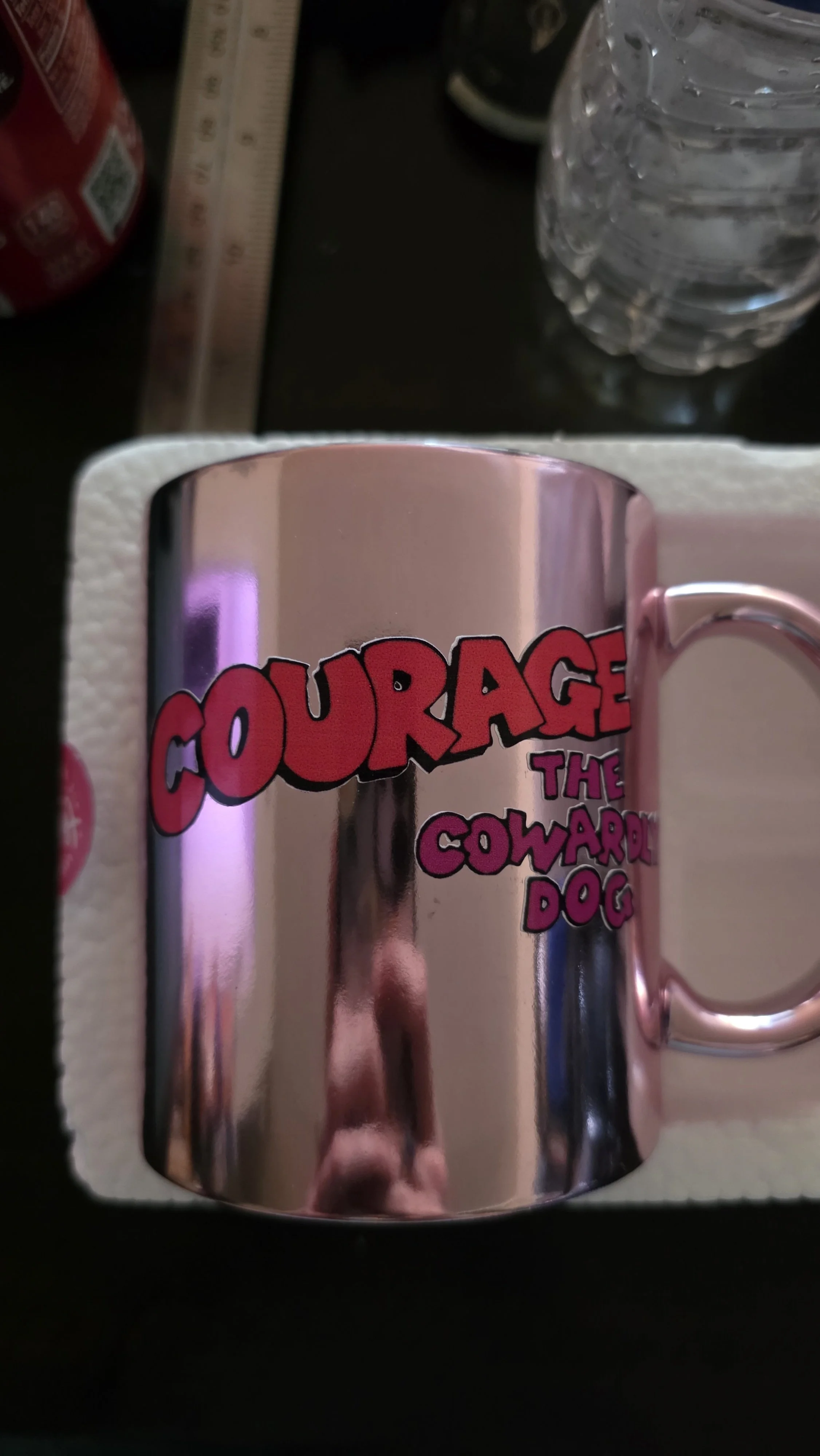 A pink mug with the words "Courage The Cowardly Dog" in red and purple letters. The mug is on a white foam tray with a bottle of water and a ruler nearby.