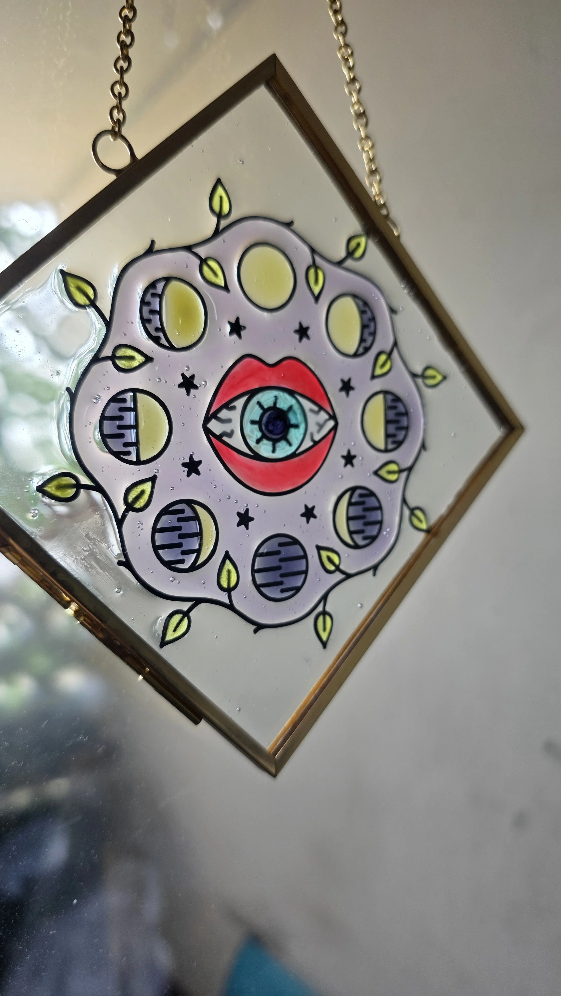 Decorative stained glass piece featuring an eye in the center with red lips, surrounded by moon phases and plant-like details, hanging from a gold chain.