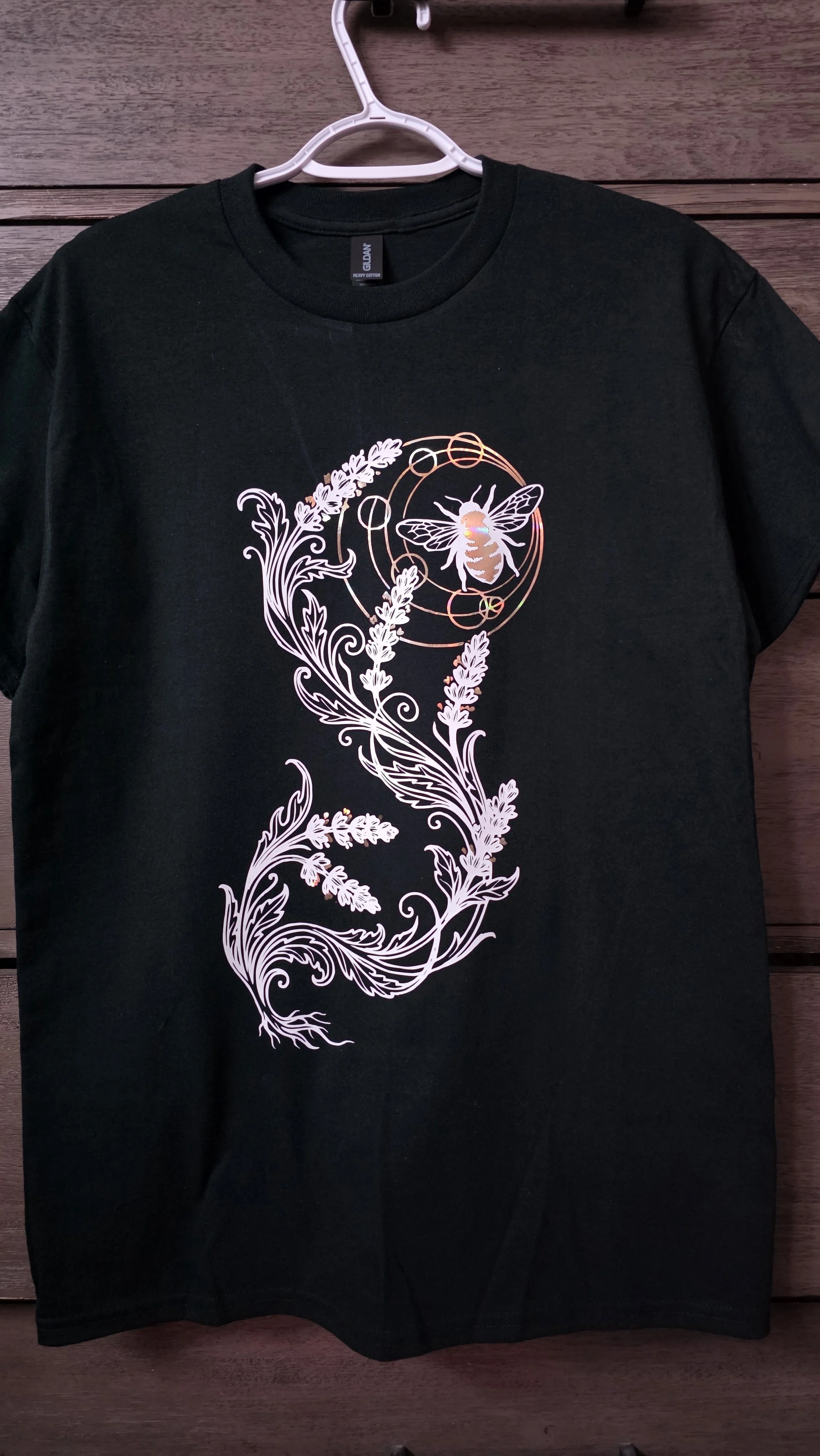 Black T-shirt with a graphic of a bee inside a circular design surrounded by floral patterns, hanging on a white hanger against a wooden background.
