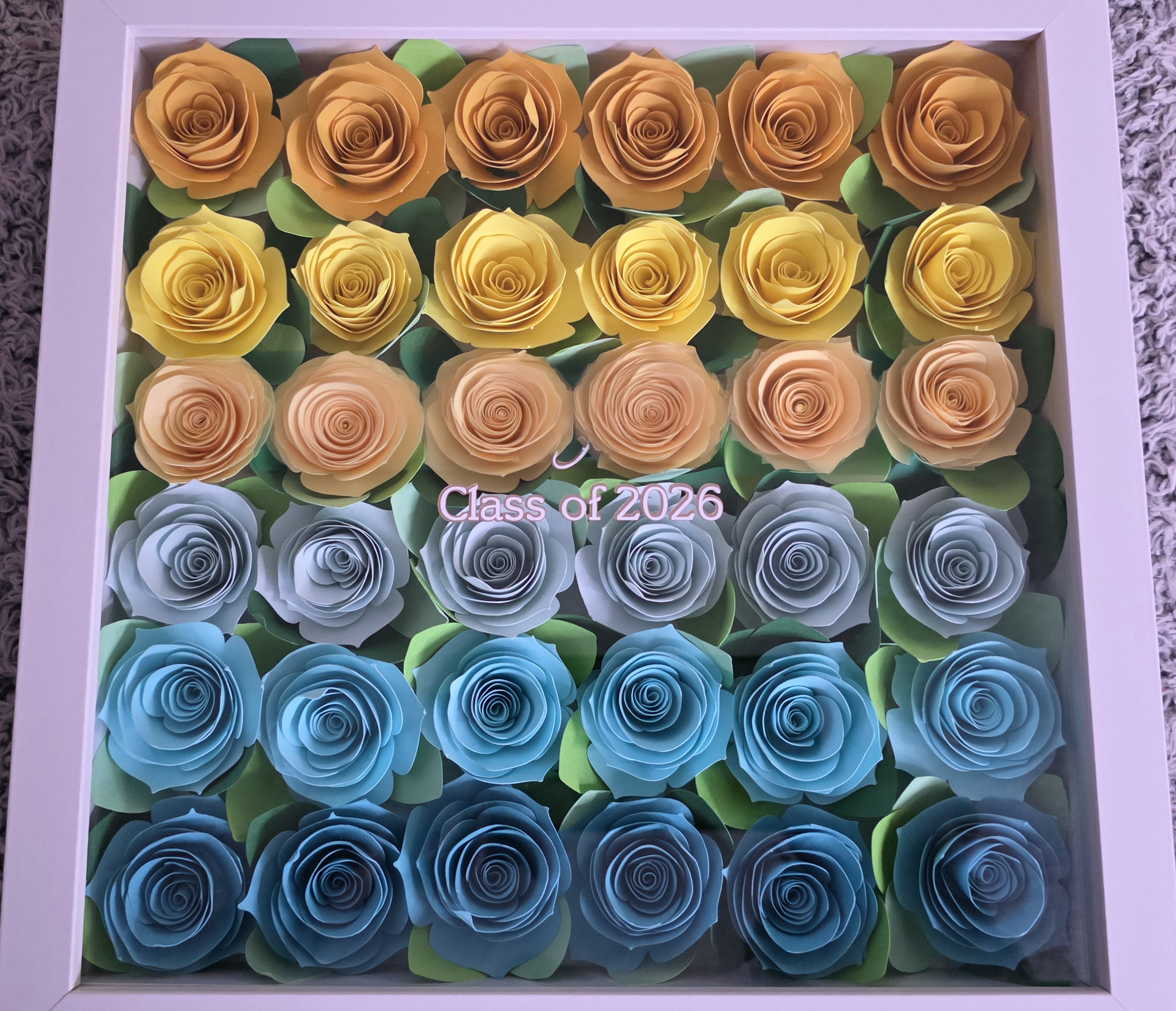 A framed display of 20 paper roses arranged in four rows, with yellow roses in the top two rows, blue roses in the bottom two rows, and green paper leaves in the middle. The frame is white and sits on a textured surface.