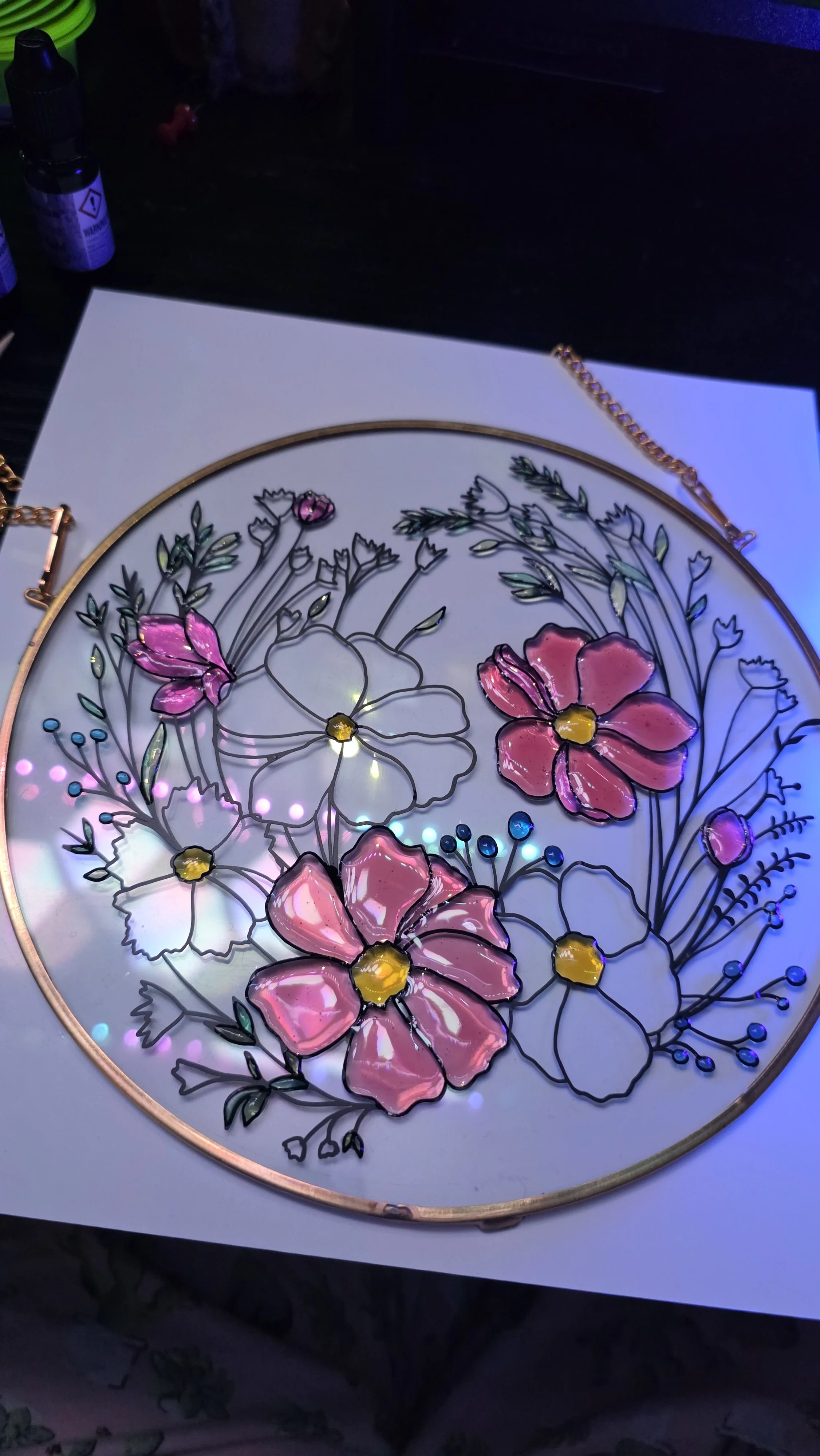 Colorful floral stained glass artwork with pink, white, and yellow flowers, illuminated by soft lights, on a white surface.
