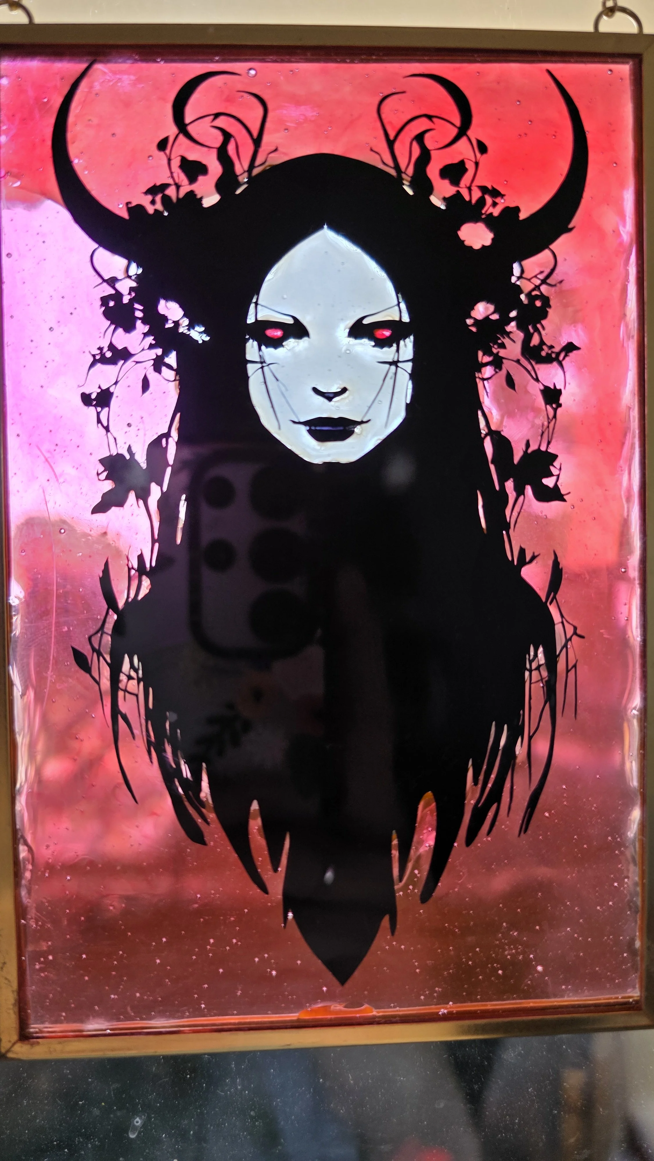 Black silhouette of a woman with long flowing hair, horns, and plant-like details, set against a pink and red background. The woman's face is centered with glowing pink eyes and dark lips.