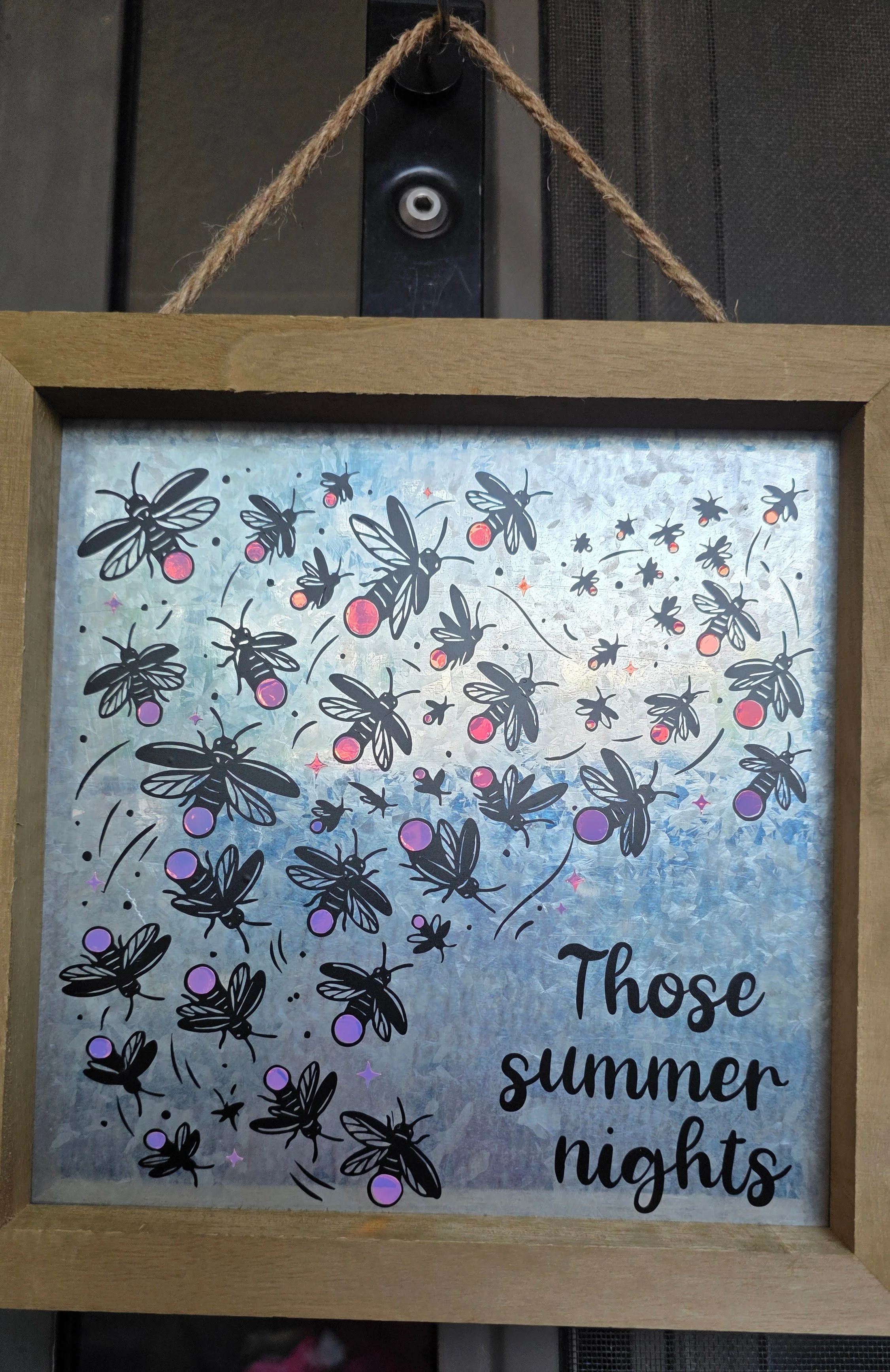 A framed decorative sign featuring a pattern of black bees with purple and pink circles for bodies. The background is a shiny, reflective silver surface, and there is partially visible black cursive text at the bottom that says 'Those summer nights'.