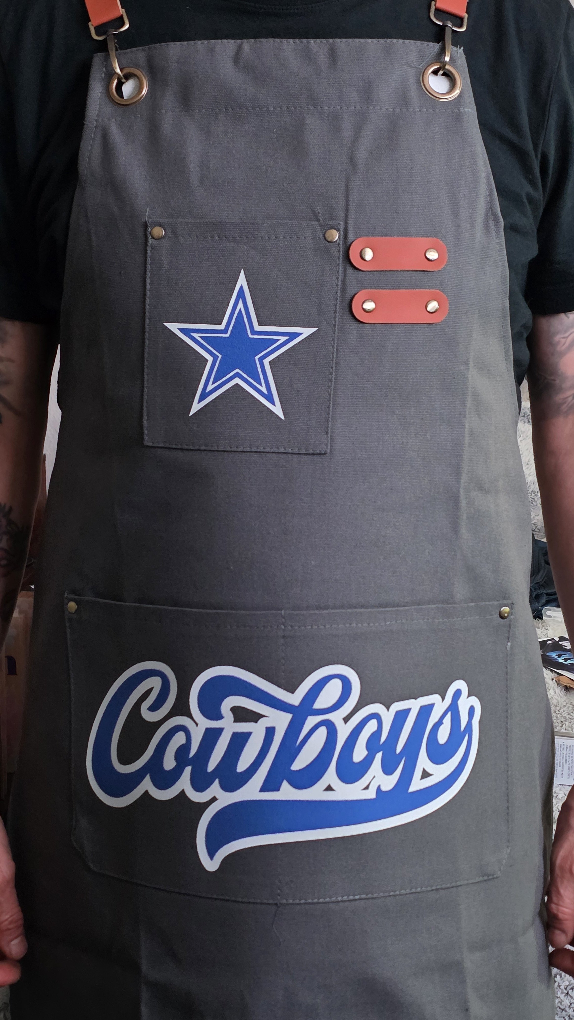 Person wearing a dark gray apron with a Dallas Cowboys star patch, red straps, and a large graphic with the word 'Couthbay' on the front.