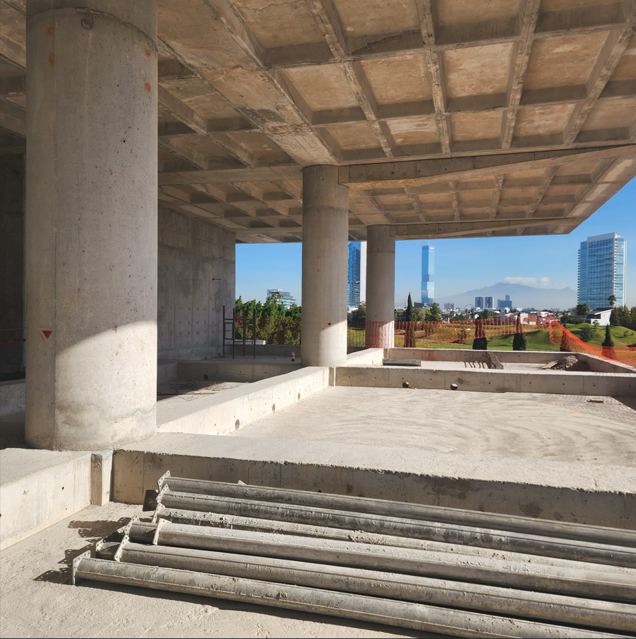 Estructura de construcción con pilares de concreto y techo de concreto en construcción, materiales metálicos en primer plano y ciudad en el fondo.
