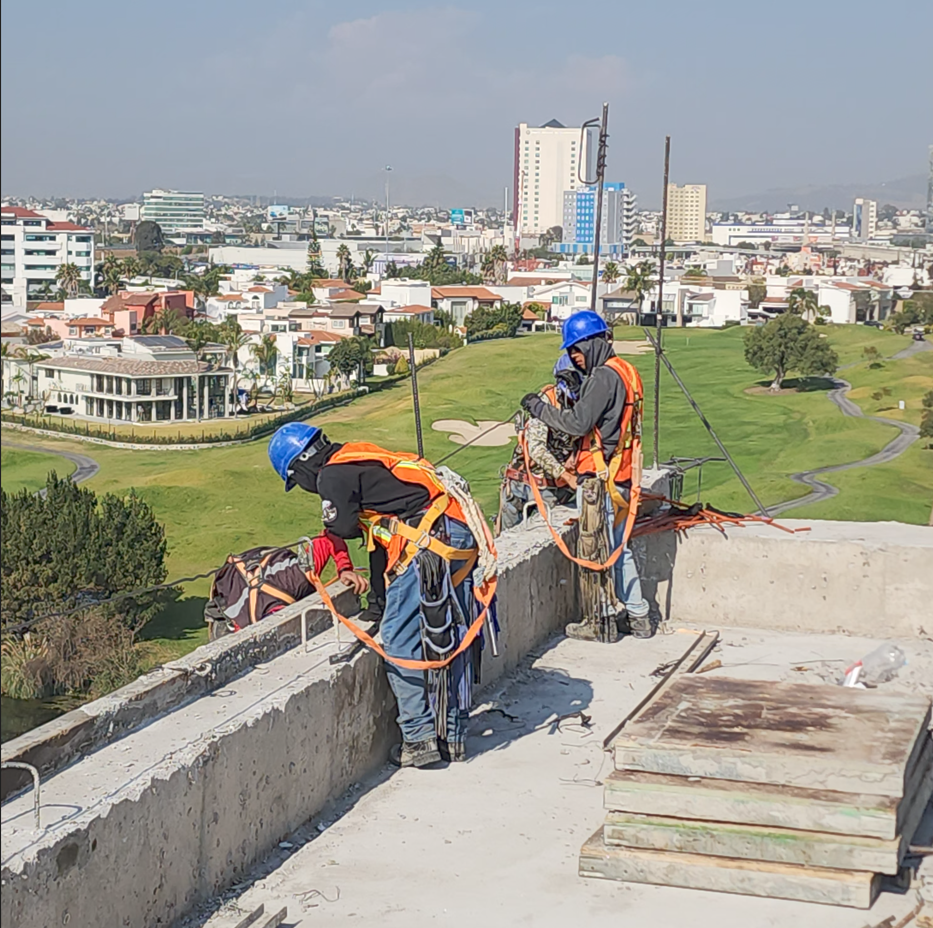 Tres trabajadores en la cima de un edificio en construcción, usando cascos y arneses de seguridad, con vistas a una ciudad y un parque verde en el fondo.