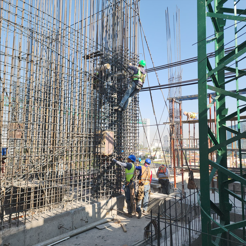 Operarios en una construcción de estructura de acero, algunos en andamios trabajando, otros en el suelo supervisando.