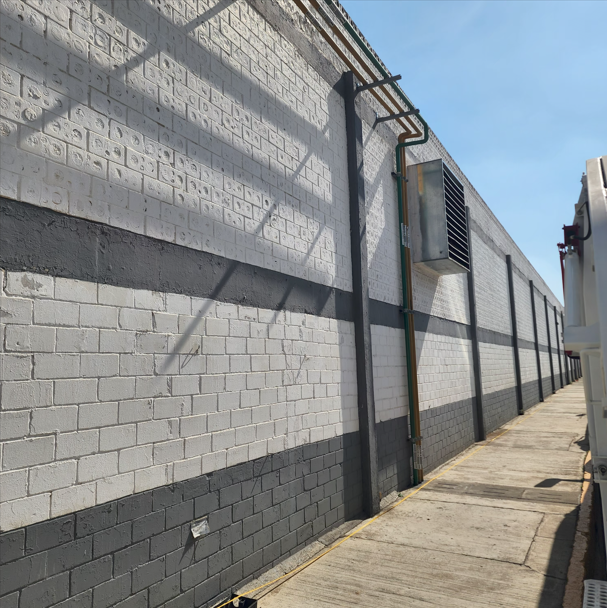 Muro de ladrillos pintados en gris, con tuberías y una unidad de aire acondicionado, junto a una acera de cemento, en un día soleado.