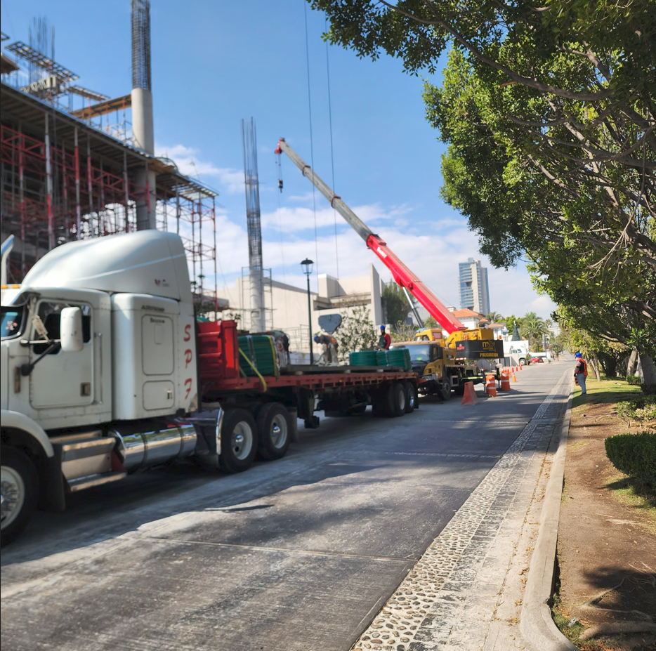 Trabajadores y maquinaria de construcción en una obra en la ciudad, con grúas y estructura en construcción, y árboles a la derecha.