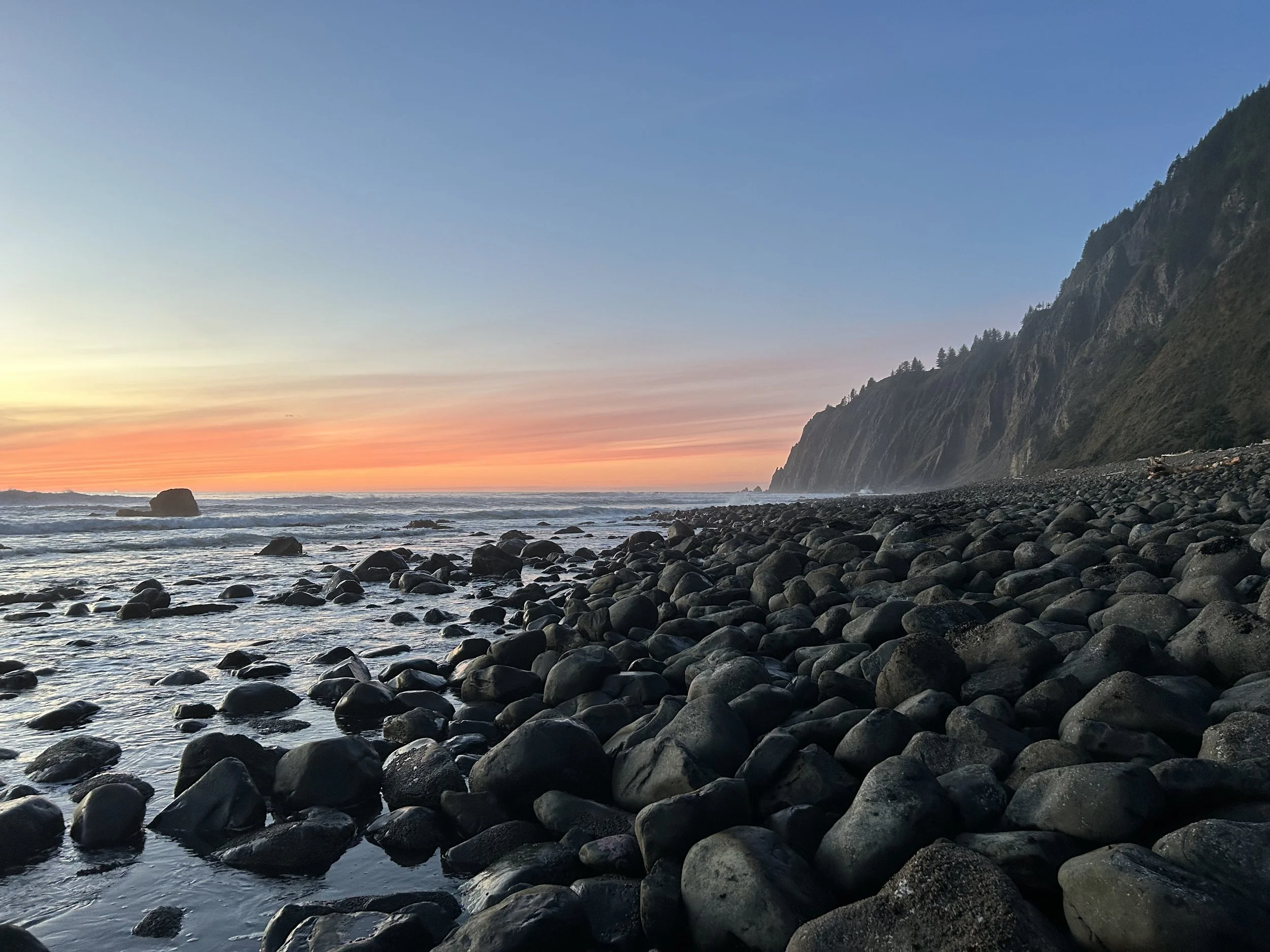 A rocky beach at sunset with waves washing over the rocks, a steep hillside covered in trees on the right, and a colorful sky with shades of orange, pink, and blue.