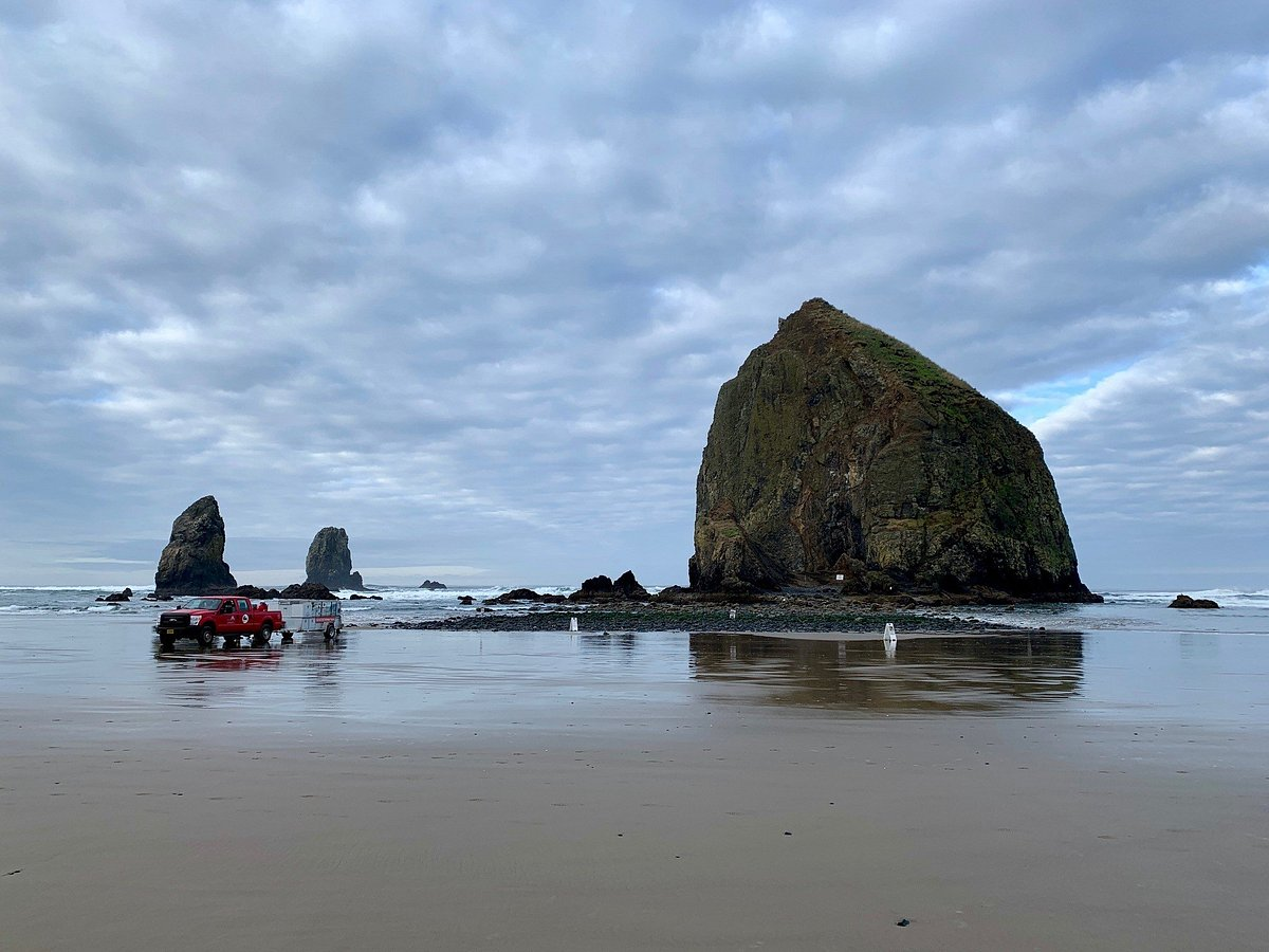 Scenic coastal view with large rock formations emerging from the ocean, wet sandy beach, cloudy sky, and a red vehicle parked near the shoreline.