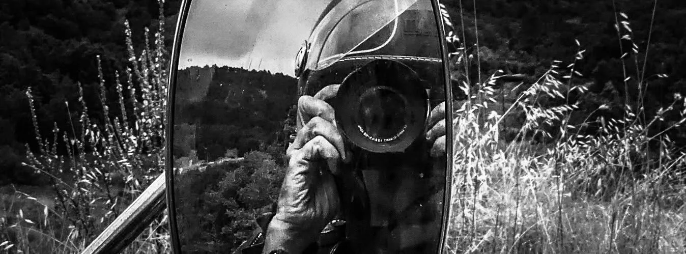 Photo en noir et blanc de l'objectif d'un appareil photo reflété dans un miroir de voiture, avec un paysage de campagne en arrière-plan.