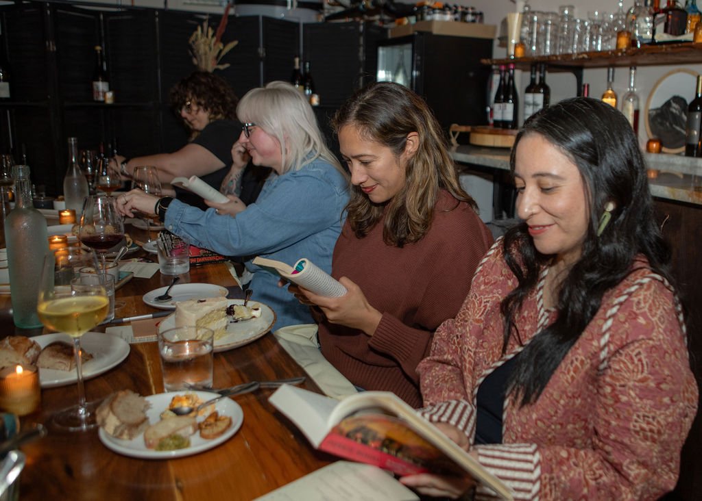 Four women sitting at a restaurant table with wine and desserts, looking at menus and books.