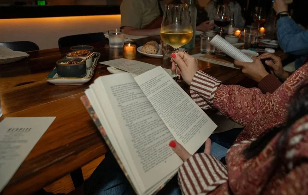 Attendees at a silent reading and wine event sit around a wooden table with open books, glasses of wine, and small shared plates, lit by candlelight.