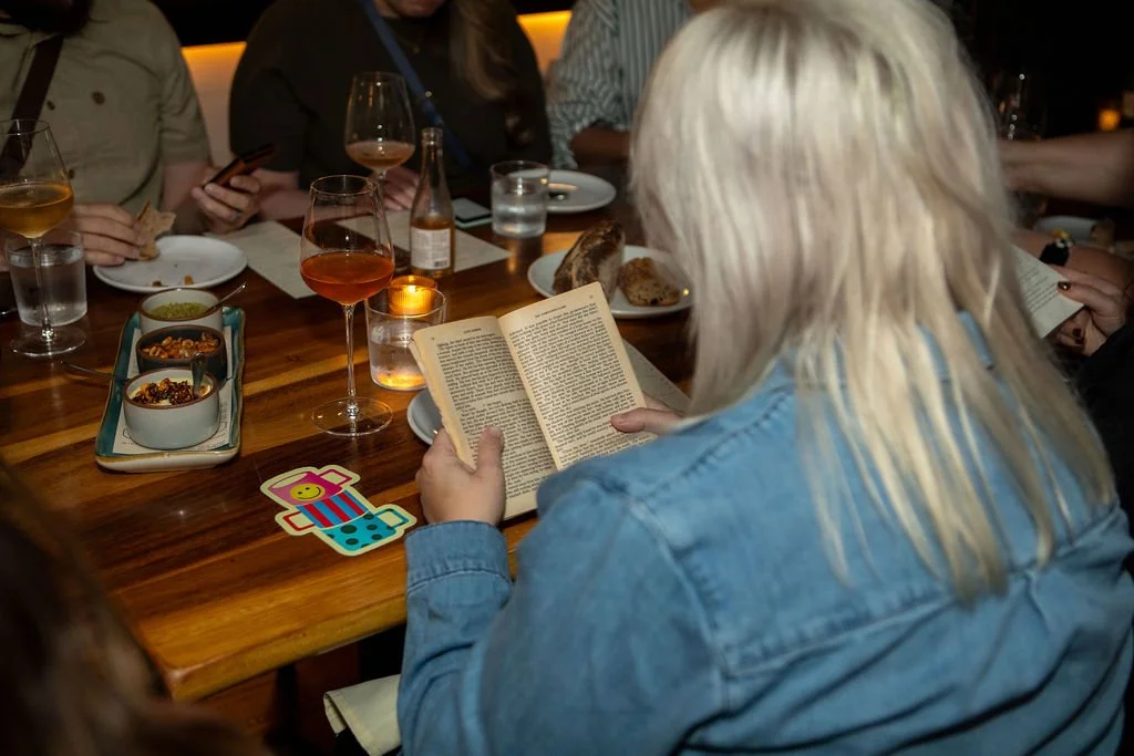 A group of people sitting around a wooden table with drinks, snacks, and a lit candle, with a woman with blonde hair reading a book.