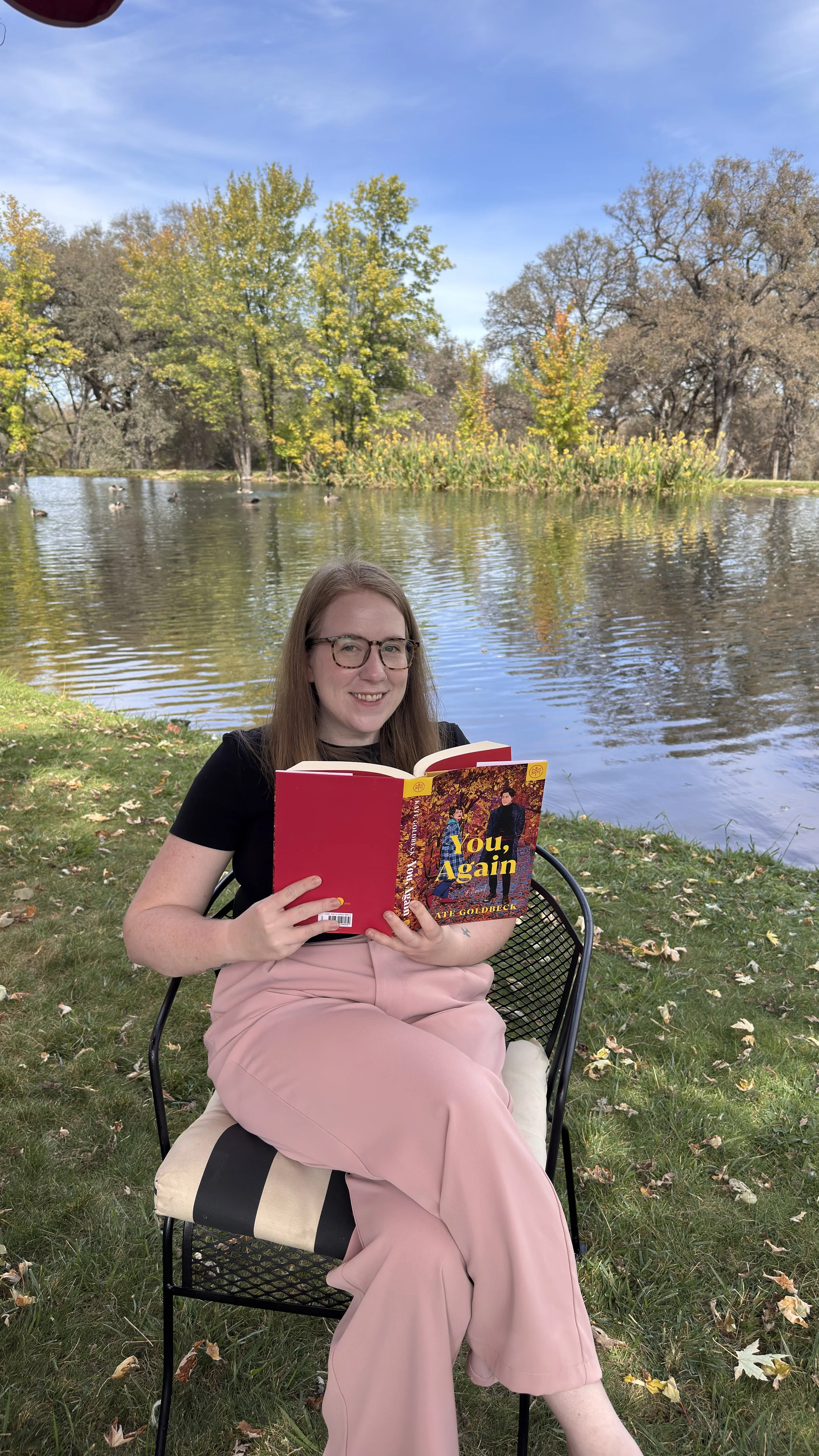 A woman with glasses and long brown hair sitting on a chair outdoors by a lake, holding a red book titled "You, Again" by Kate Goldbeck, with trees and ducks in the background on a clear day.