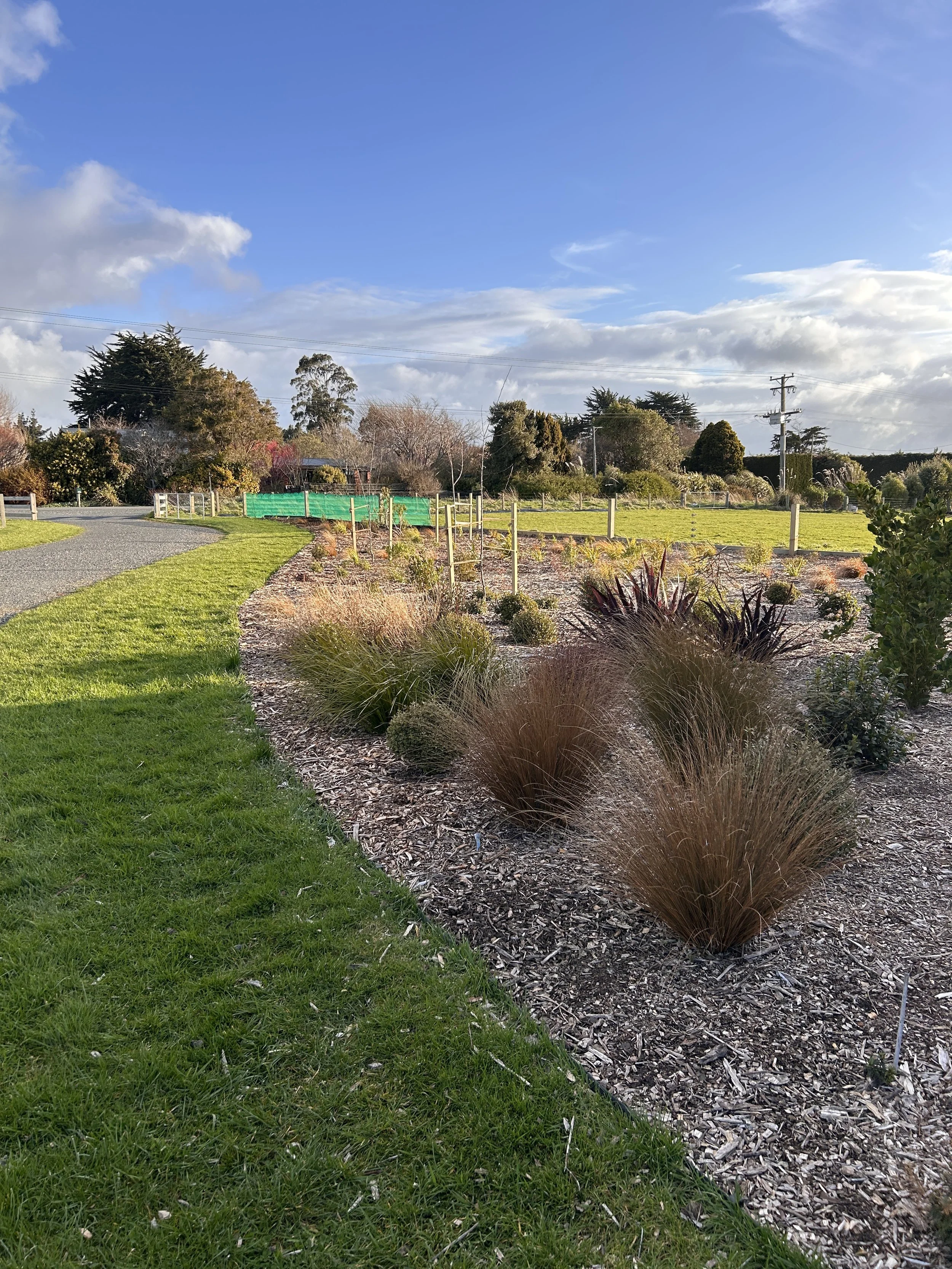 A landscaped garden with a curved gravel pathway, green grass, and decorative plants. In the background, there are trees, bushes, and power lines under a partly cloudy blue sky.