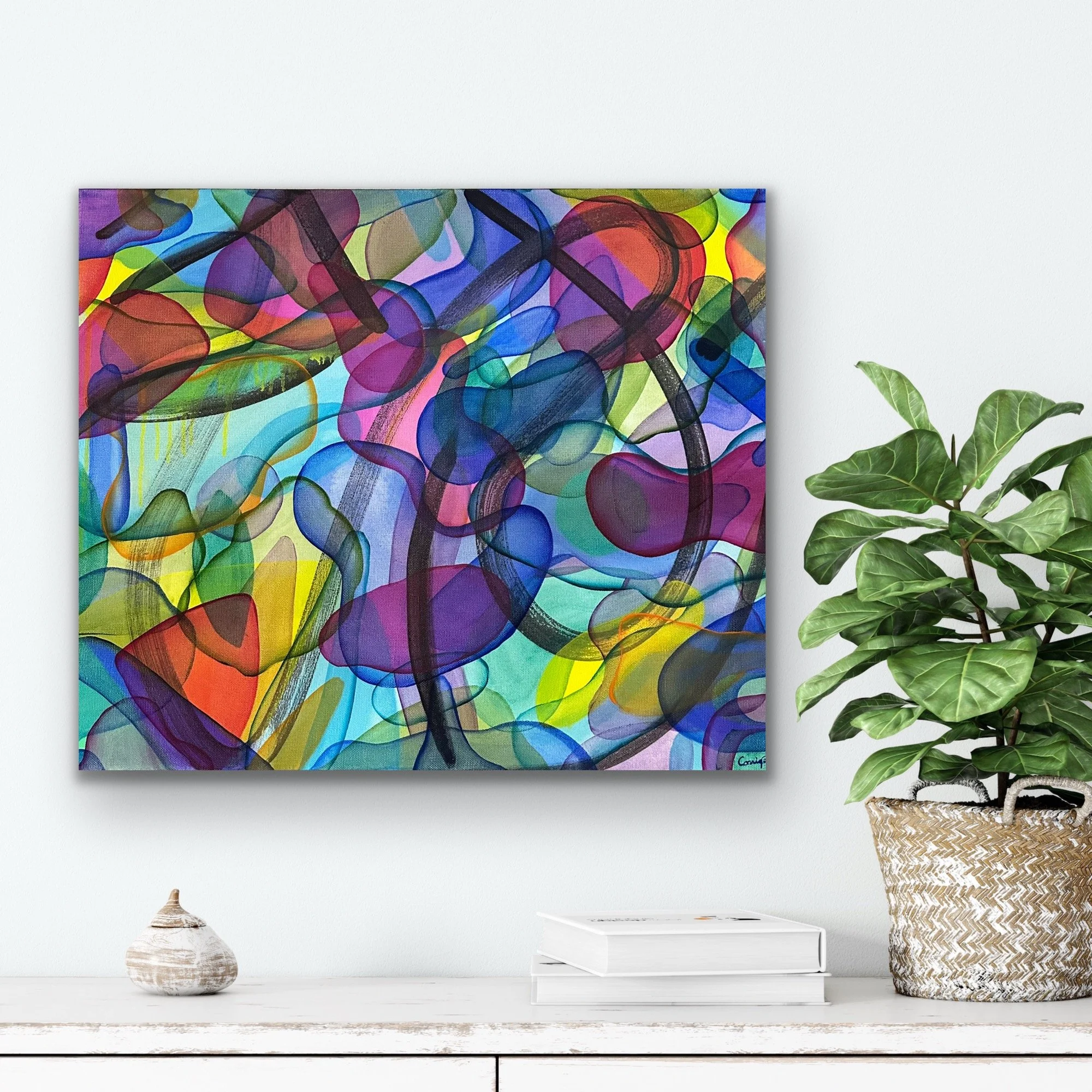 A colorful abstract painting hanging on a white wall above a white desk with books, a ceramic vase, and a potted plant in a wicker basket.