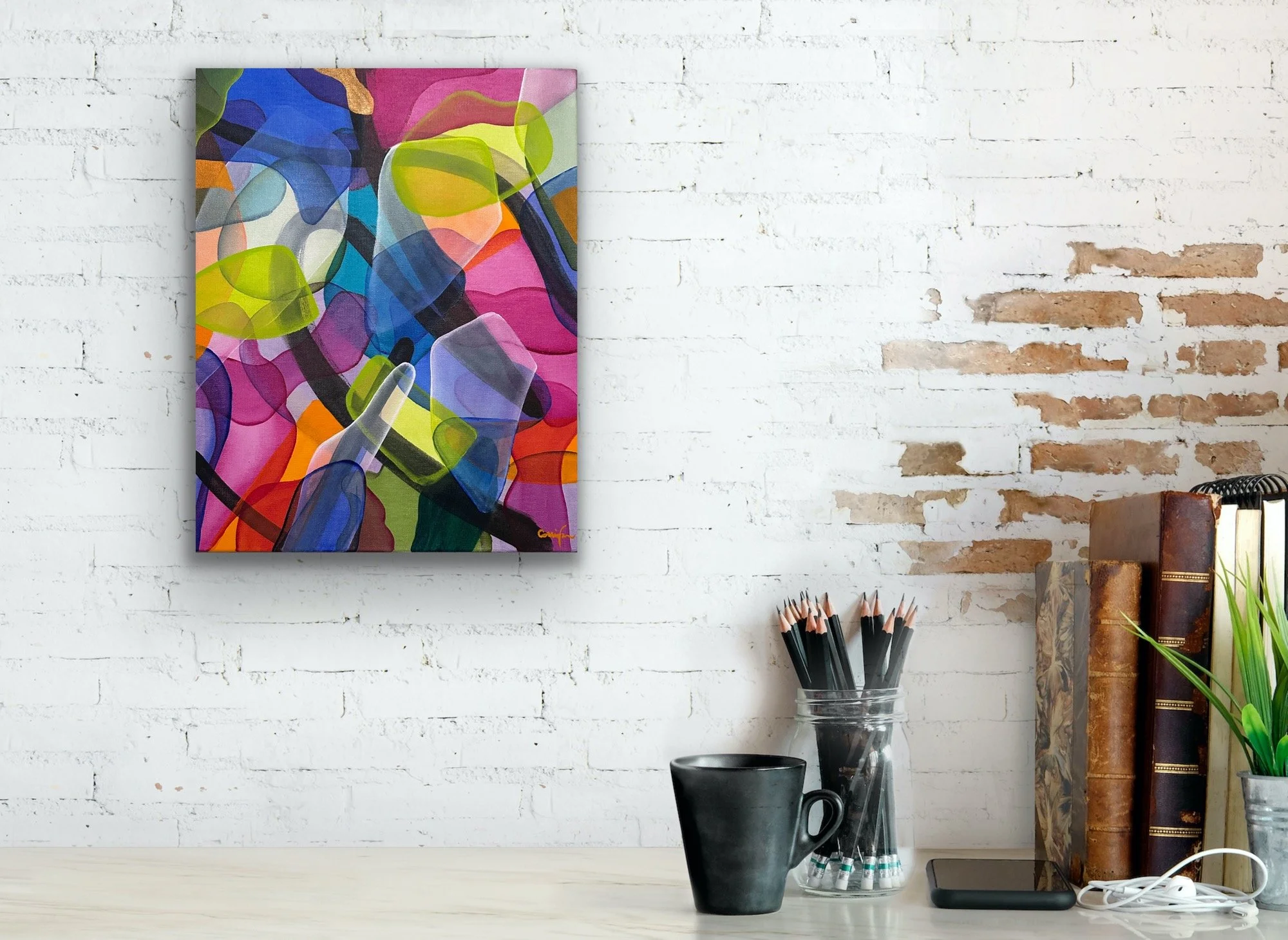 A colorful abstract painting hangs on a white brick wall. On the desk beneath it, there is a glass cup filled with black pencils, a black mug, a smartphone, and a pair of white earphones. To the right, a stack of books with decorative covers and a po