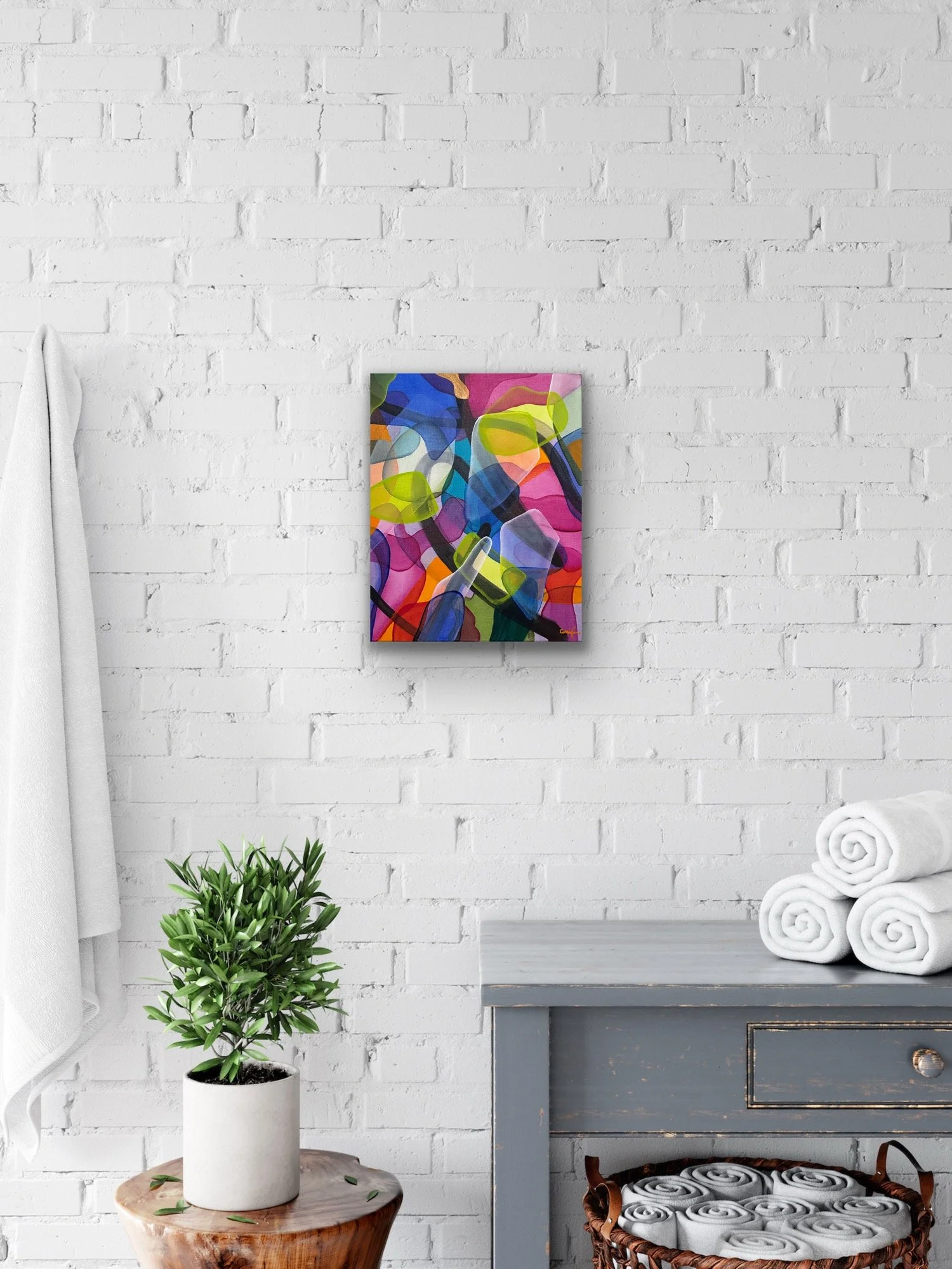 A decorated room corner with a white brick wall, a colorful abstract painting, a potted green plant on a wooden table, a gray wooden piece of furniture with rolled white towels and a basket with rolled towels on top.