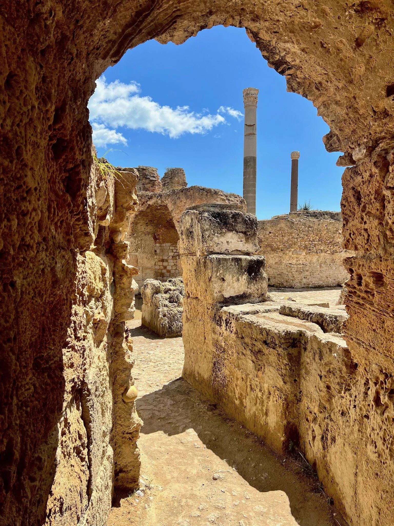 Ancient ruins viewed through a stone archway with tall columns in the background against a blue sky with a few clouds.