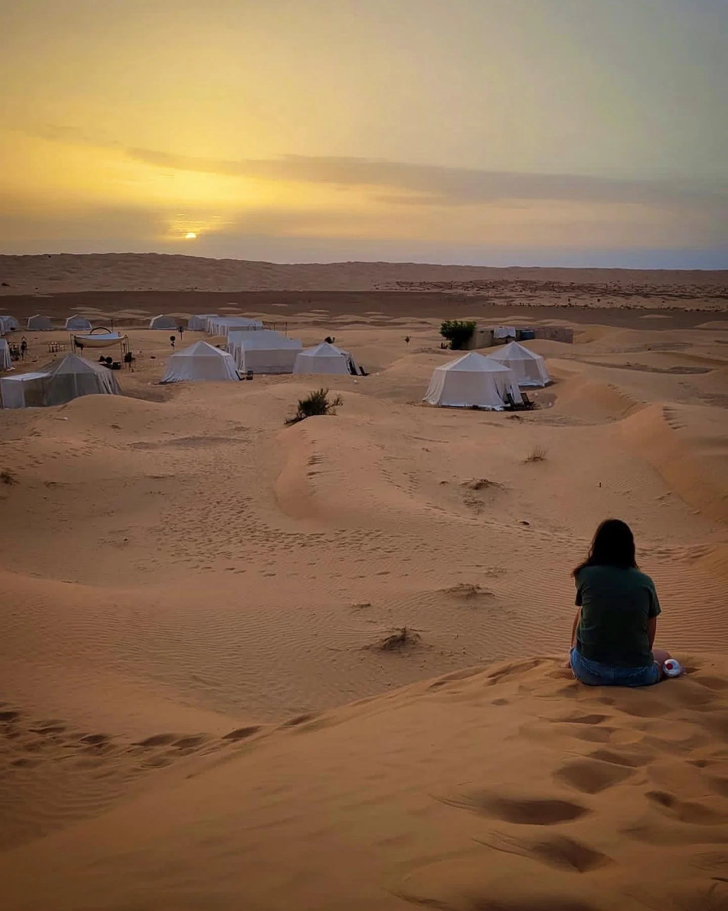 Person sitting on sandy dunes watching a desert camp with white tents during sunset.
