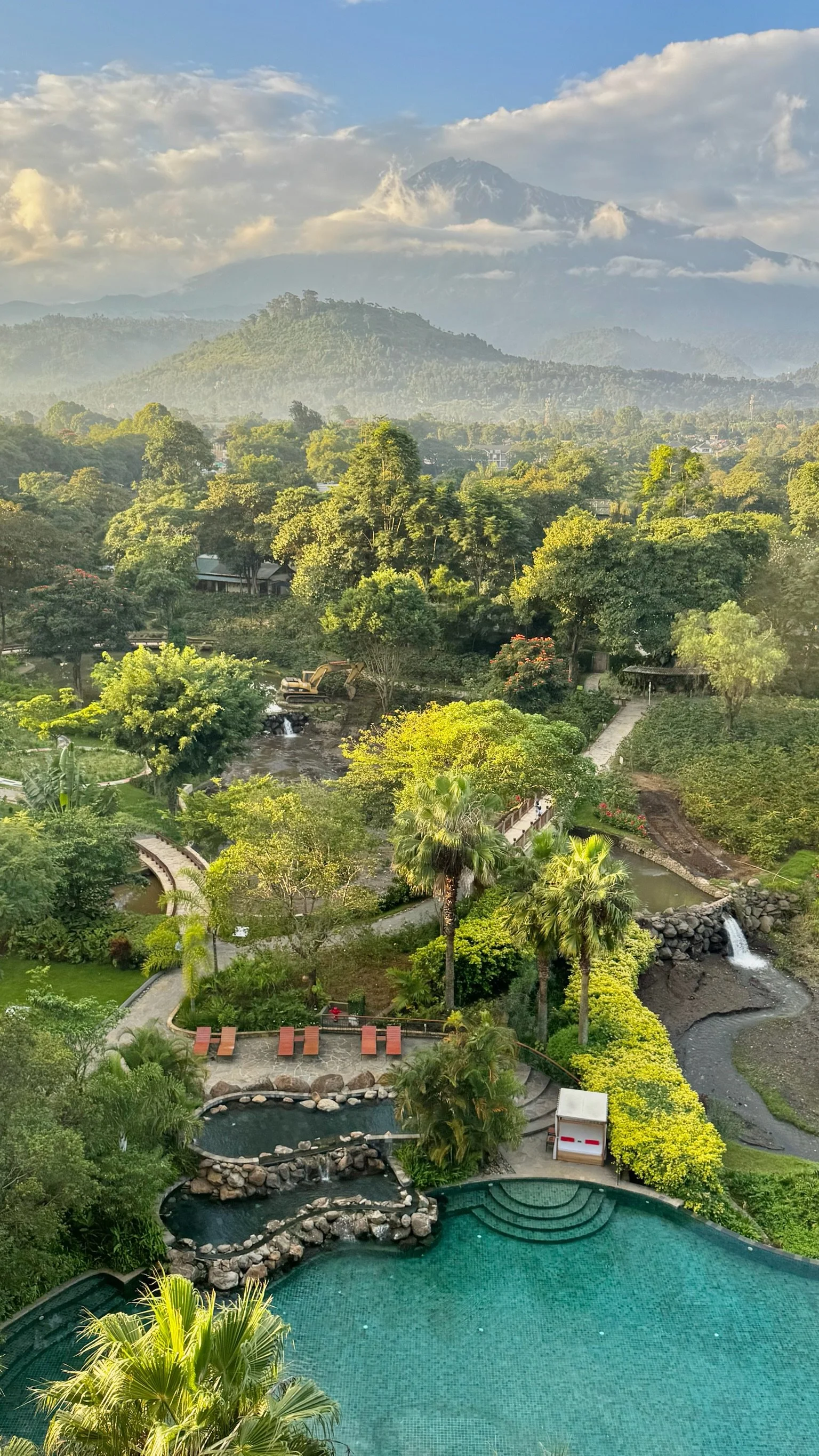 Scenic view of a lush, green landscape with multiple pools, waterfalls, and walkways, surrounded by trees, with mountains in the background and partly cloudy skies.