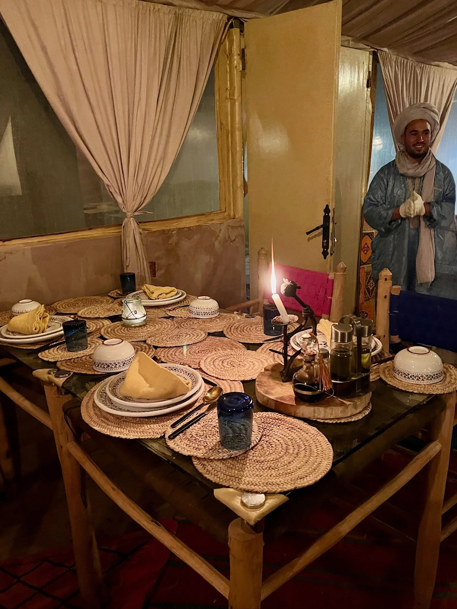 A dining table set with woven placemats, white bowls, black drinking glasses, yellow cloth napkins, and silverware. The table is under warm lighting, with a lit candle and a candle holder in the foreground.