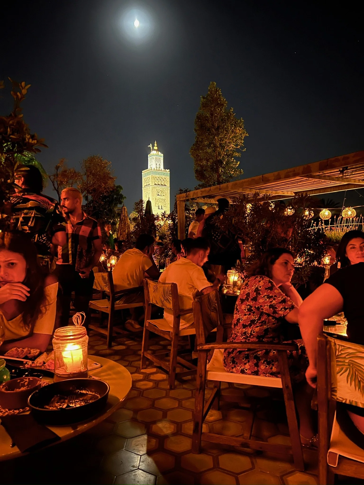 Nighttime scene at a rooftop restaurant with people dining, warm candlelight, and the moon shining in a dark sky. The Leaning Tower of Pisa is visible in the background.