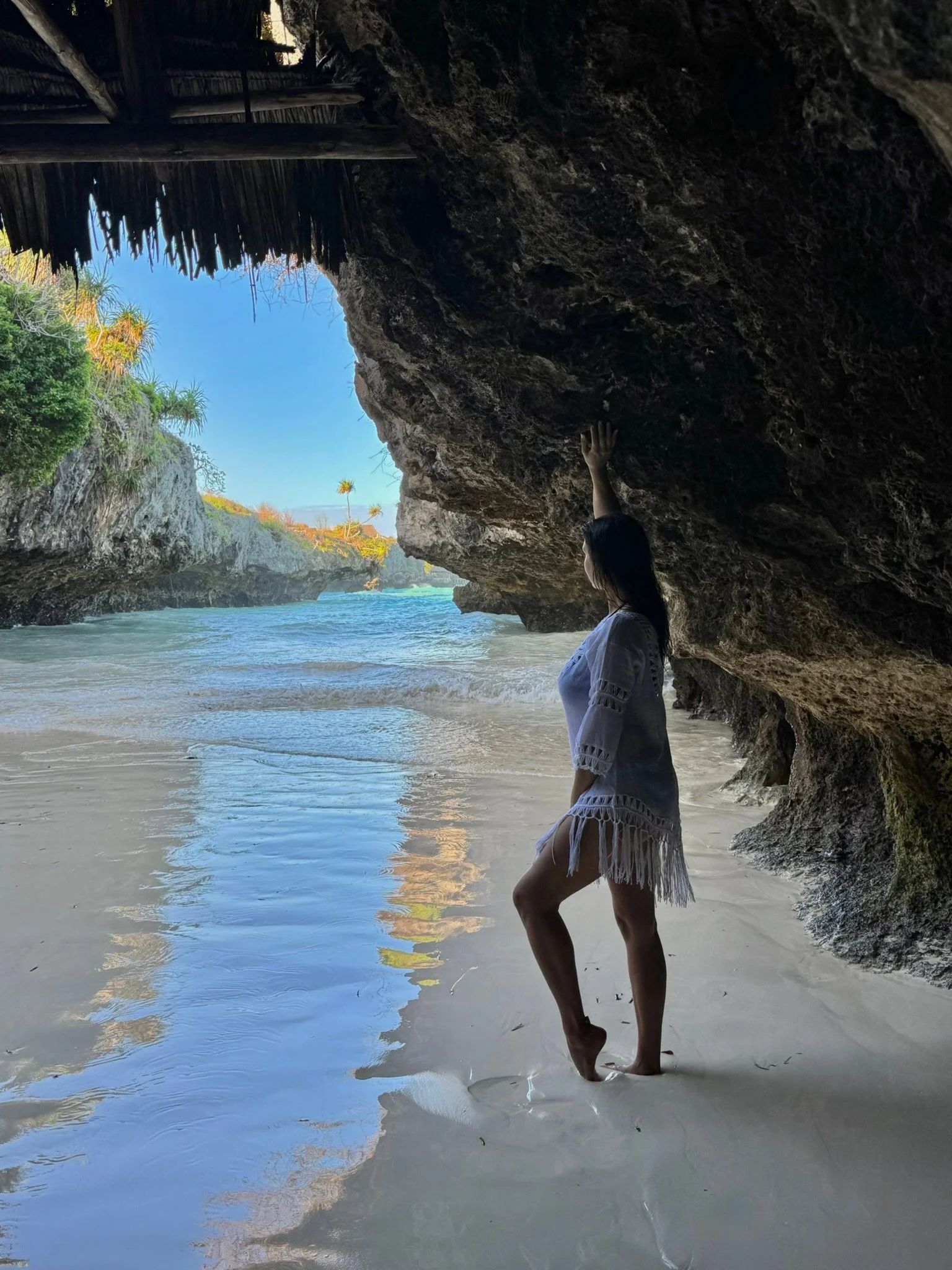 Woman standing under a rocky overhang on a beach, touching the ceiling of the cave with her hand, with ocean water and a blue sky visible in the background.