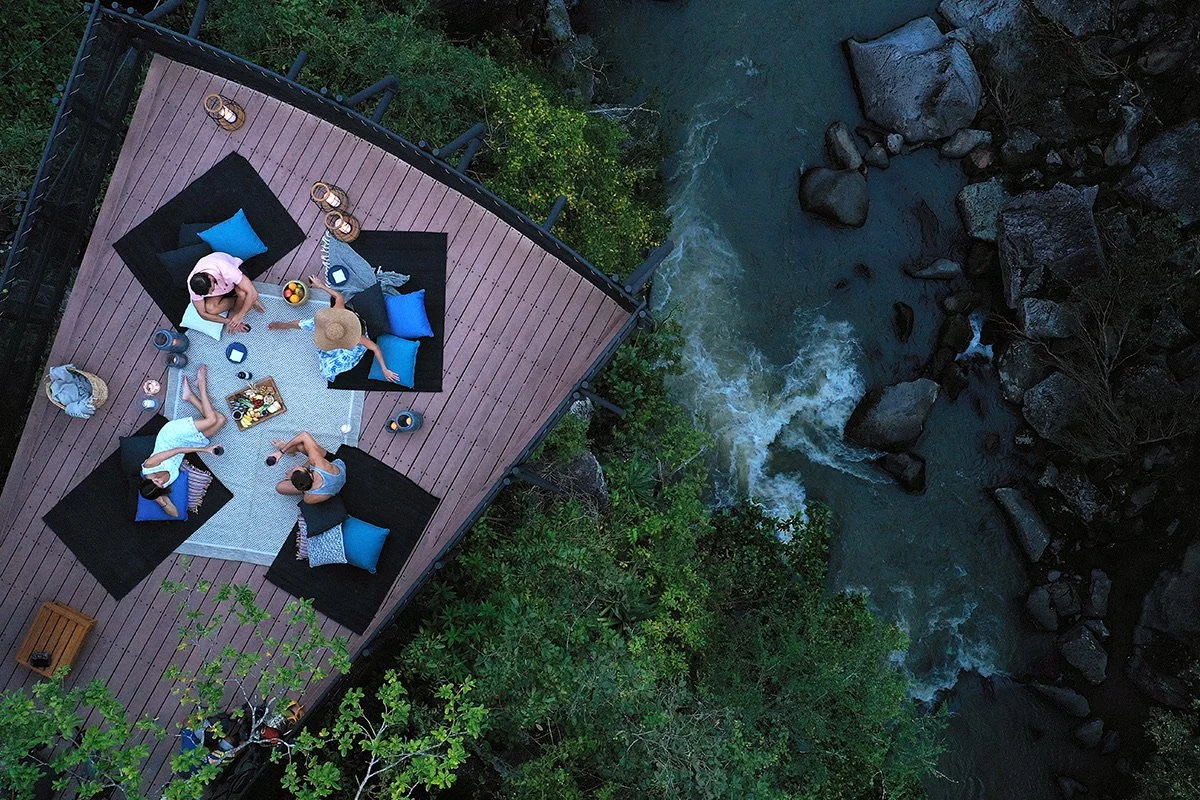 Aerial view of four people having a picnic on a wooden deck next to a flowing river surrounded by trees
