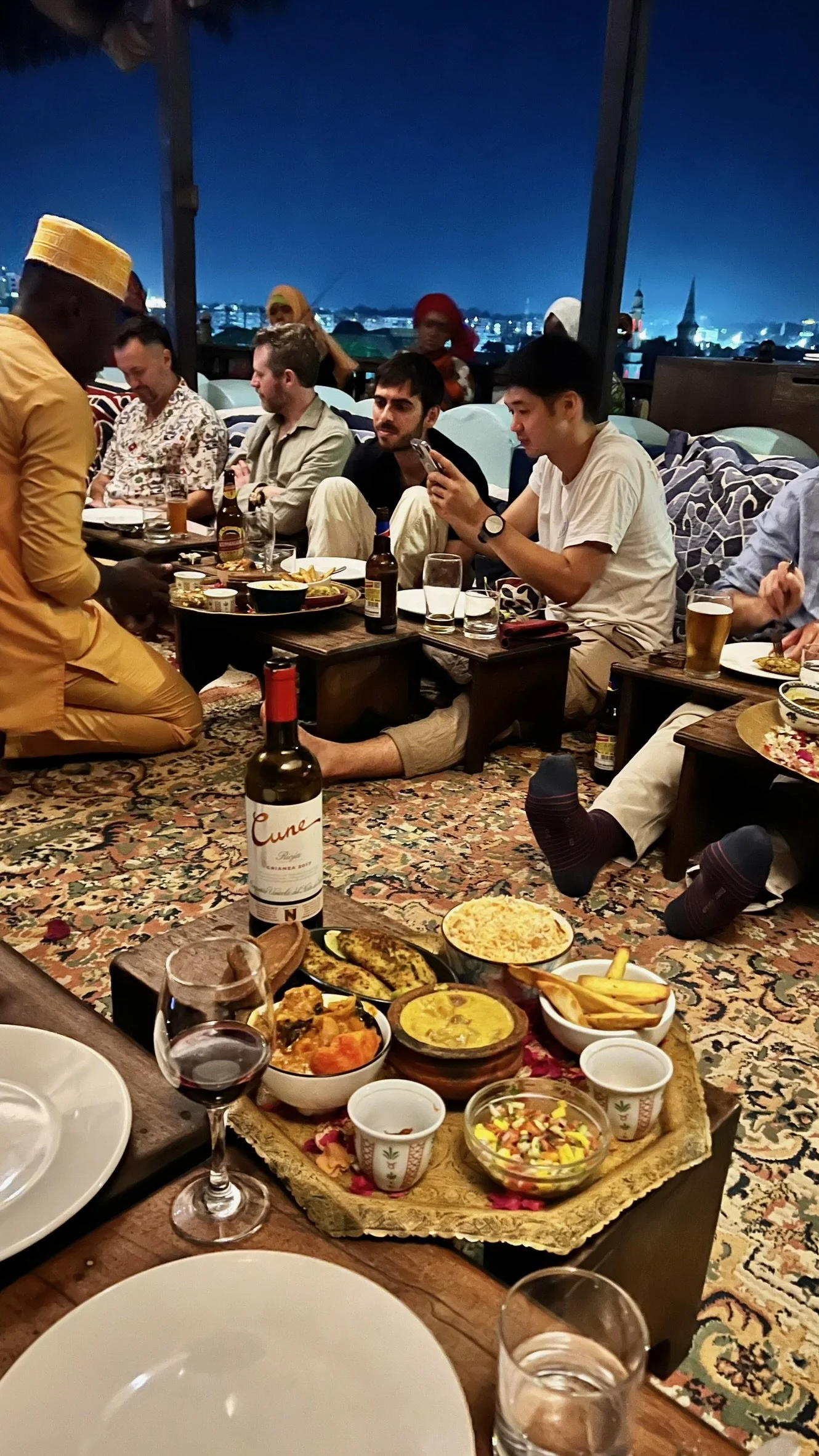 A group of people sitting on the floor around a low table filled with food, drinks, and family-style dishes. The setting appears to be indoors during the evening, with city lights visible through large windows.