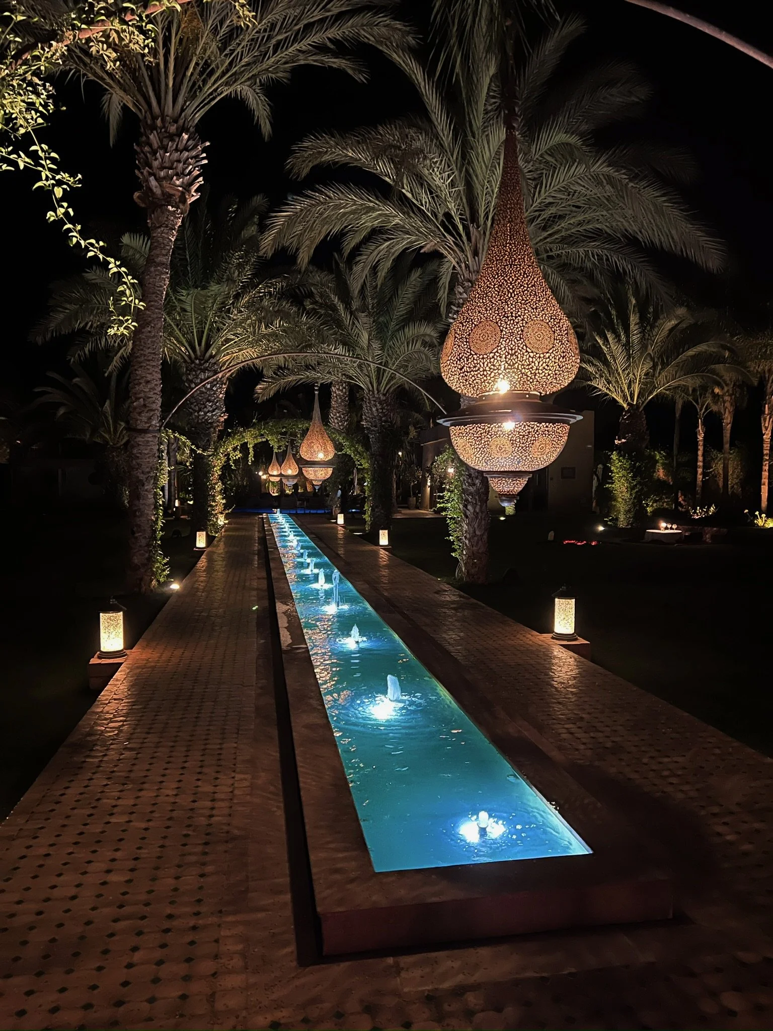 Nighttime scene with illuminated hanging lanterns, palm trees, and a water feature with small fountains in the center.