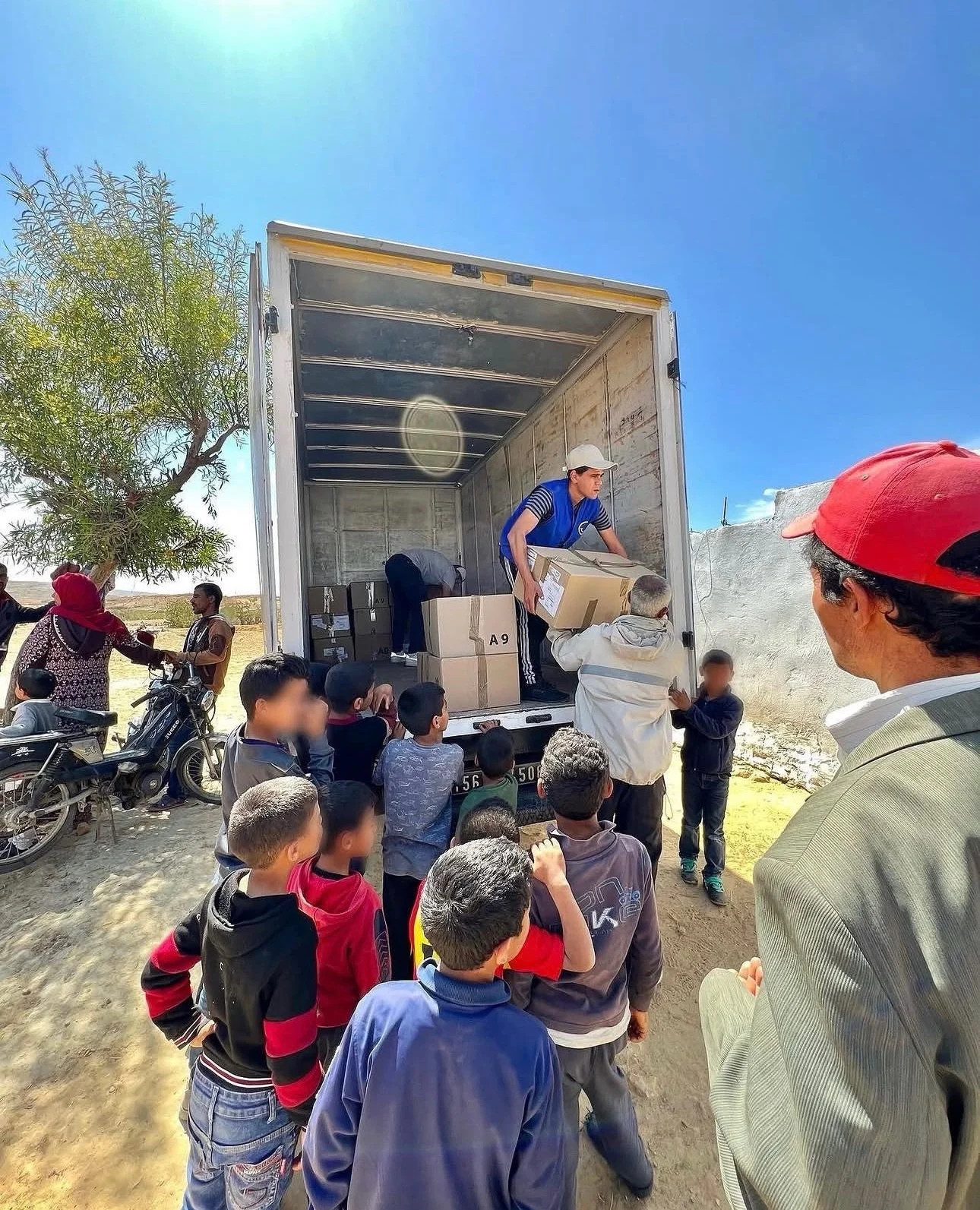 People gather around an open truck where volunteers are handing out boxes, with children and adults waiting in line outside, in an outdoor setting on a sunny day.